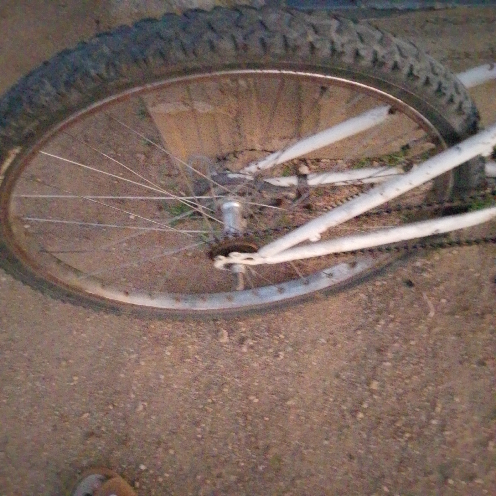 The image shows a close-up of a bicycle wheel with a knobby tire, spokes, and rim. The bicycle frame, painted white, is partially visible. The wheel is on the ground, which appears to be a dirt or gravel surface. The background is out of focus but suggests an outdoor environment. A person's shoe is visible at the bottom left corner of the image. The scene is set in Wuro Allahamdu, Nigeria.