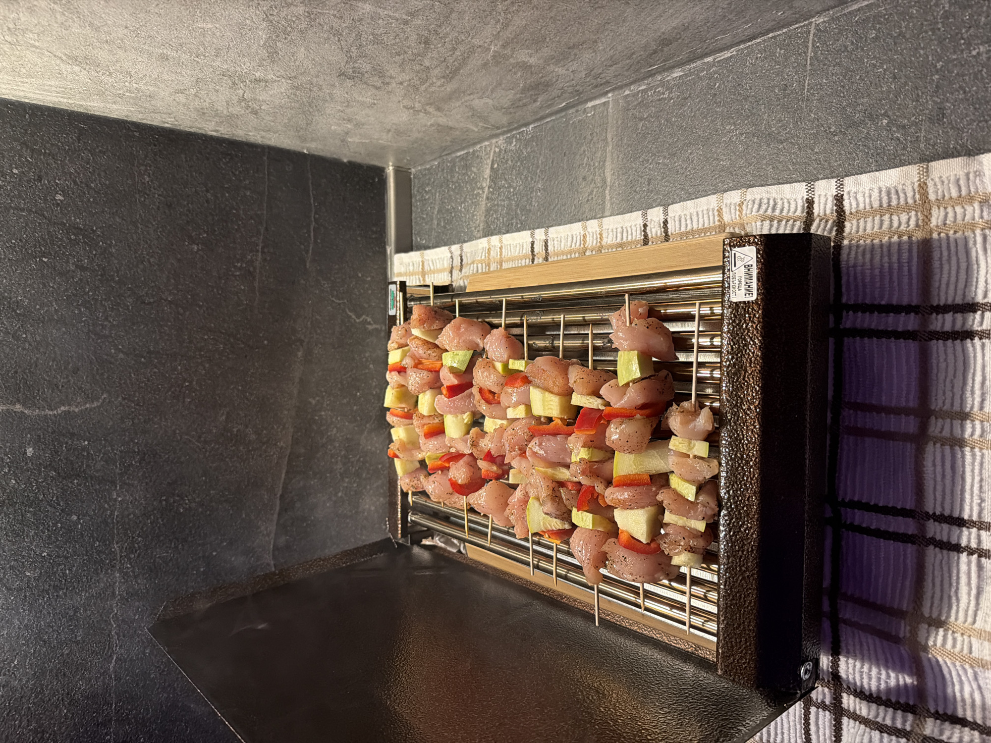 The image shows a close-up, high-angle view of a portable electric grill with several skewers of raw chicken, red bell peppers, and zucchini on its grate. The grill is open, and the skewers are arranged in rows. The grill itself is dark brown and sits on a checkered cloth. The background consists of a dark, textured wall. There are no people or animals present. The scene appears to be indoors, possibly in a kitchen or on a covered balcony, given the texture of the wall and the presence of a countertop or similar surface. The lighting suggests artificial illumination. There is no discernible connection to the city of Plovdiv, Bulgaria.