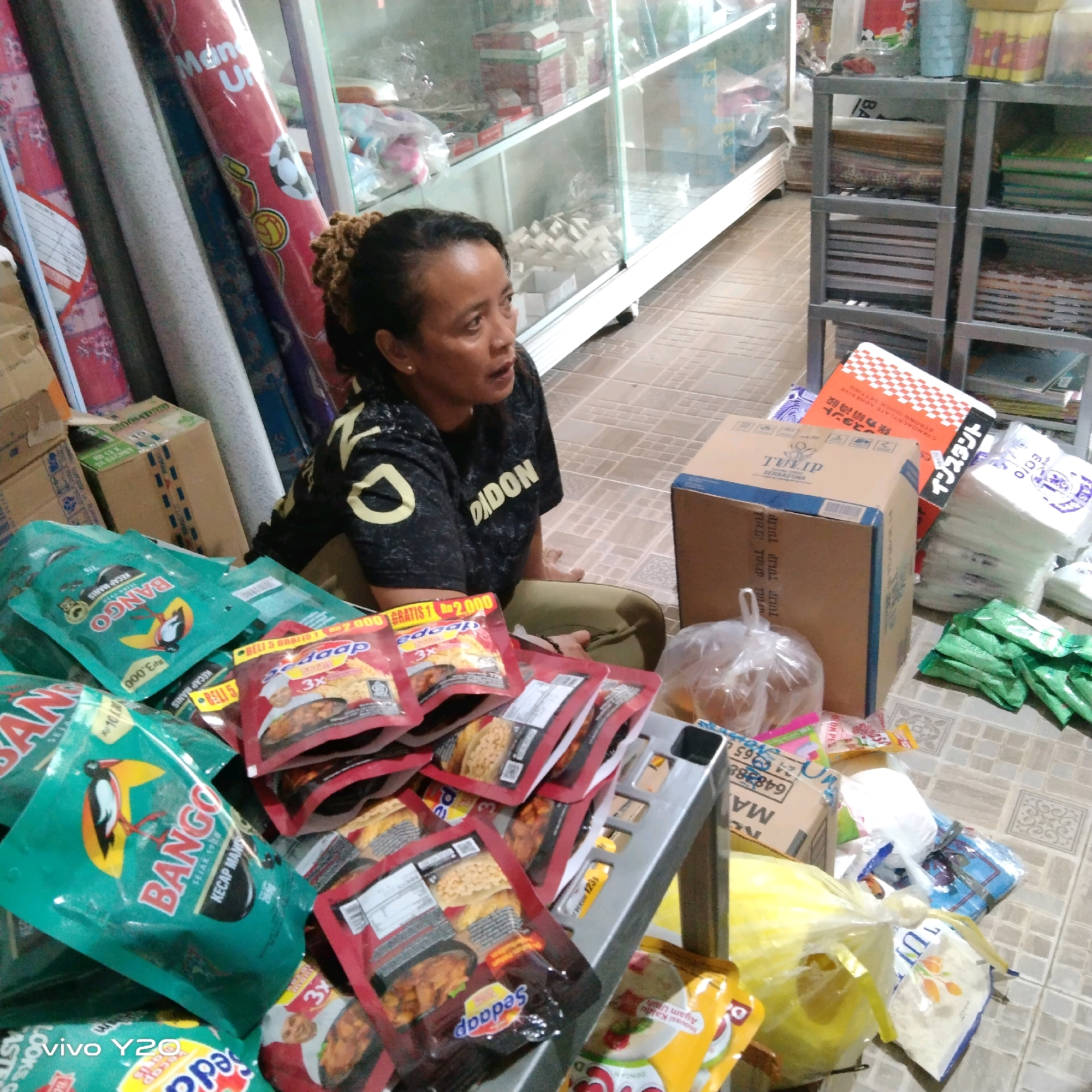 The image depicts an indoor scene, possibly a store or storage area, in Unknown, Unknown. A woman with dark, curly hair is seated on the floor, looking to her right and speaking. She is wearing a dark t-shirt with yellow markings on the sleeves and front, and olive-green pants. The floor is tiled and somewhat dirty.
Surrounding the woman are numerous products. To her left, there are several large, rolled-up items that appear to be carpets or fabric, some with decorative patterns. In front of her, piled on the floor and a gray metal shelf unit, are many packaged food items, mostly in red and green pouches. Prominent brands include "BANGO" (in green packaging) and "Sedap" (in red packaging), suggesting these are condiments or sauces. There are also several cardboard boxes, one of which has the brand "TULIP" printed on it.
Other objects visible include plastic bags filled with items, stacks of white paper products or tissues, and more assorted goods on shelves in the background. The overall impression is one of a retail or distribution environment with a substantial quantity of goods.