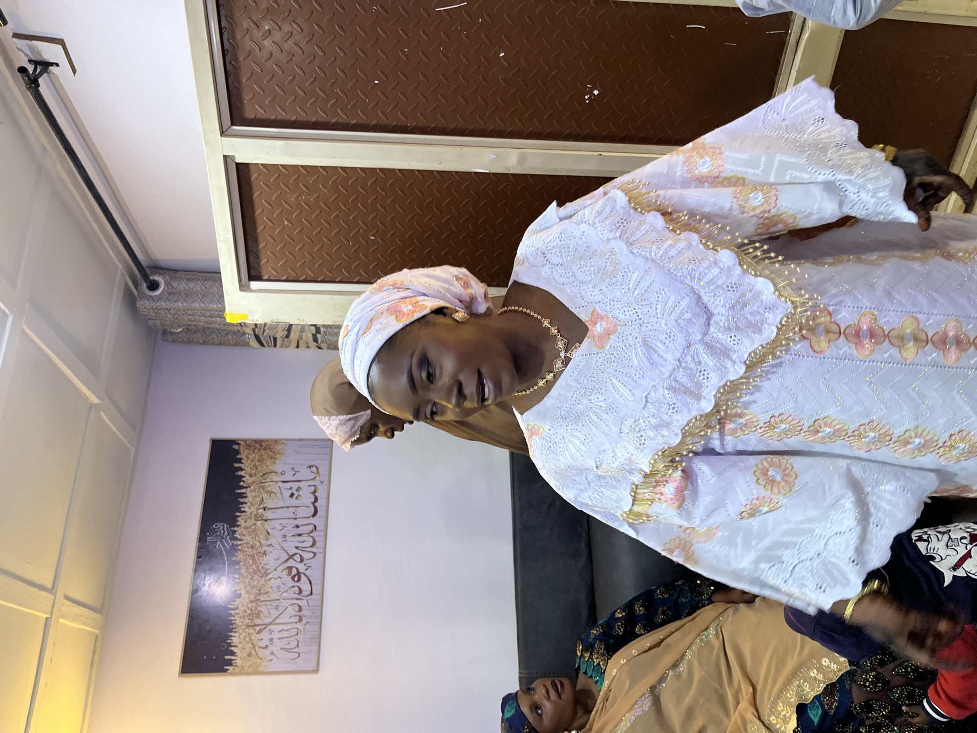 The image depicts an indoor scene, likely a home, in the city of Jos, Nigeria. In the foreground, a woman is prominently featured, facing left and speaking, wearing a white, intricately embroidered dress with gold and pink floral patterns and gold trim. She also wears a pink headscarf and gold jewelry, including a necklace and earrings. To her left, two other women are seated on a dark sofa. One, closer to the foreground, wears a tan and blue patterned outfit and a blue headscarf. The other, slightly further back and to the left, wears a tan garment with patterned blue and black detailing on her sleeve. Behind the main subject, another woman, partially visible, is wearing a brown garment and a light pink headscarf. The background includes a white wall adorned with a framed piece of art featuring Arabic calligraphy and a gold abstract design. A brown, textured metal door or partition is visible on the right side of the image. The ceiling appears to be made of white tiles with a grid pattern.