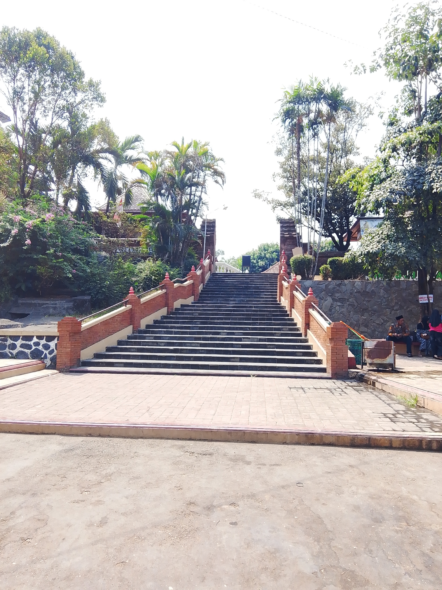 The image shows a wide, long set of stone stairs leading upwards towards a building and trees. The stairs are flanked by brick railings with decorative finials on top of the posts. To the left of the stairs is a stone wall and lush greenery. To the right of the stairs, a stone wall forms a backdrop for a seating area where two people are sitting, one wearing a traditional Indonesian outfit. The ground in front of the stairs is paved with reddish-brown bricks, transitioning to a dirt or gravel area at the bottom of the frame. The environment appears to be outdoors in a tropical or subtropical location, with tall trees and palm trees visible. The scene is brightly lit by sunlight, suggesting it is daytime. The building at the top of the stairs has a traditional architectural style with a tiled roof. The image captures a public space, possibly a temple or a historical site, in Mantingan, Indonesia.