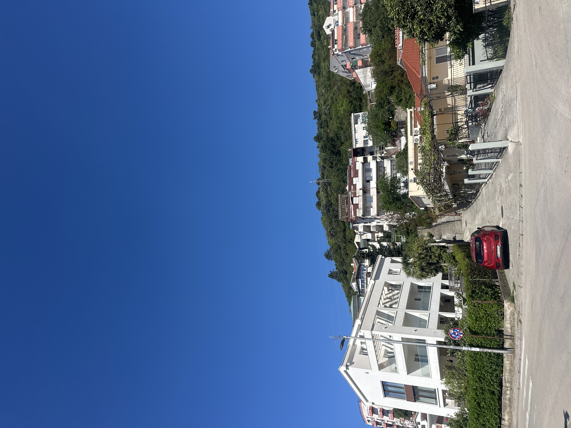 The image displays a sunny, residential street in Budva, Montenegro, characterized by a clear blue sky and lush green vegetation. The scene is dominated by a collection of multi-story buildings, primarily white or light-colored, featuring a mix of modern architecture with glass balconies and traditional styles with red-tiled roofs. These structures are built along what appears to be a hillside, surrounded by abundant trees and shrubs.
A paved road or driveway occupies the foreground on the right, where a bright red hatchback car is parked. The car's license plate, "BD 232 PJ," is clearly visible, with "BD" being the regional identifier for Budva. Adjacent to the parked car, a street light pole stands, to which a circular traffic sign, likely indicating "No Stopping" or "No Parking" (a blue circle with a red diagonal line), is attached.
The sky, occupying a significant portion of the left and upper frame, is a vibrant, cloudless blue, suggesting excellent weather conditions. Thin, almost imperceptible lines or wires stretch across the mid-ground against the sky. The bright, direct sunlight indicates that the photo was taken during the day, likely mid-morning to late afternoon, creating well-defined shadows. No people are visible in the tranquil scene. The architecture and Mediterranean-style greenery align with the provided location context of Budva, Montenegro.