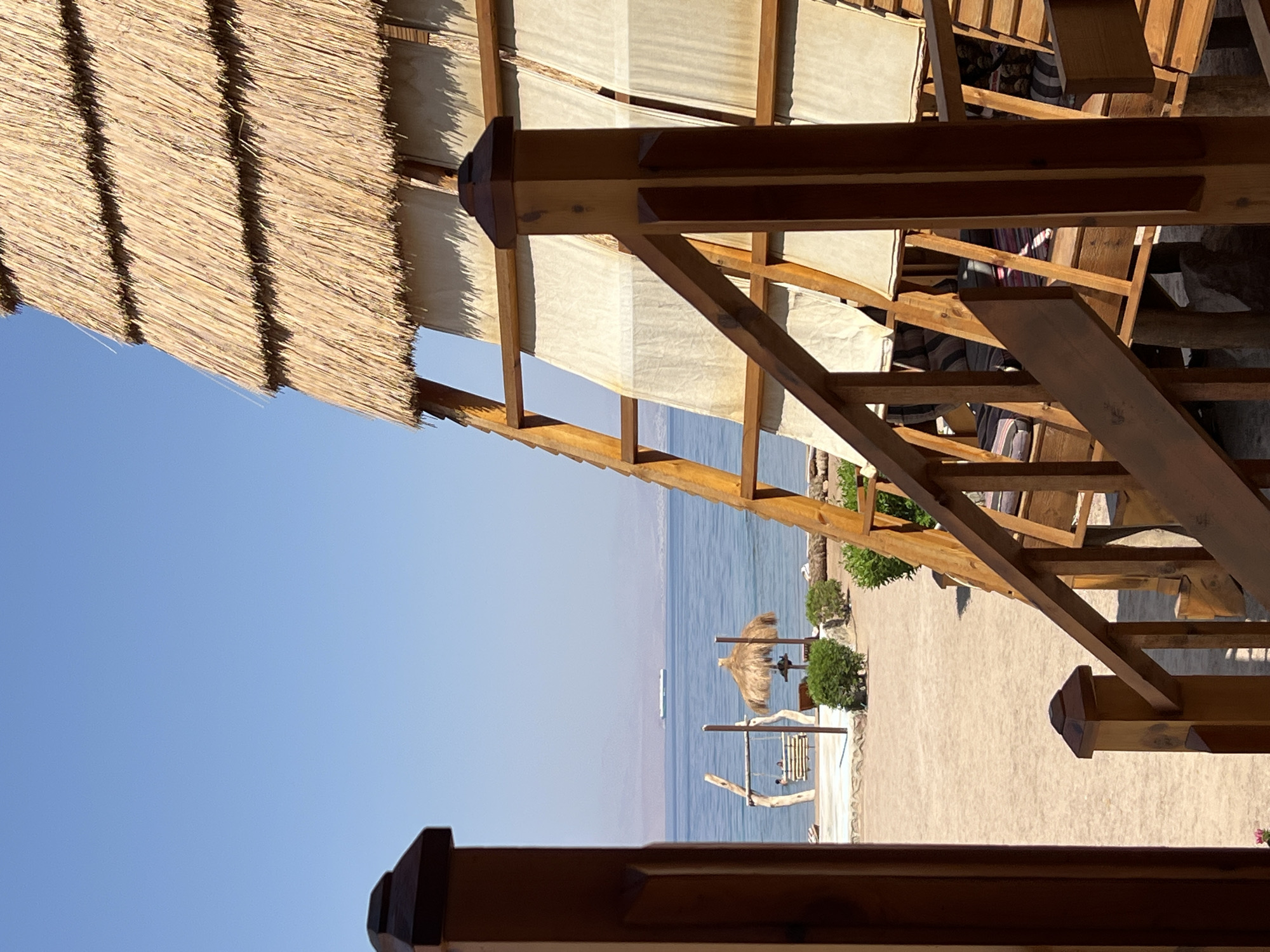 The image depicts a beachfront view in Nueiba, Egypt, featuring a thatched-roof structure with wooden railings and steps in the foreground. Beyond the structure, a sandy beach leads to a calm blue sea under a clear sky. A distant cargo ship is visible on the horizon. A wooden swing with two people sitting on it and a thatched umbrella are situated on the beach. The scene suggests a tranquil resort or coastal setting.