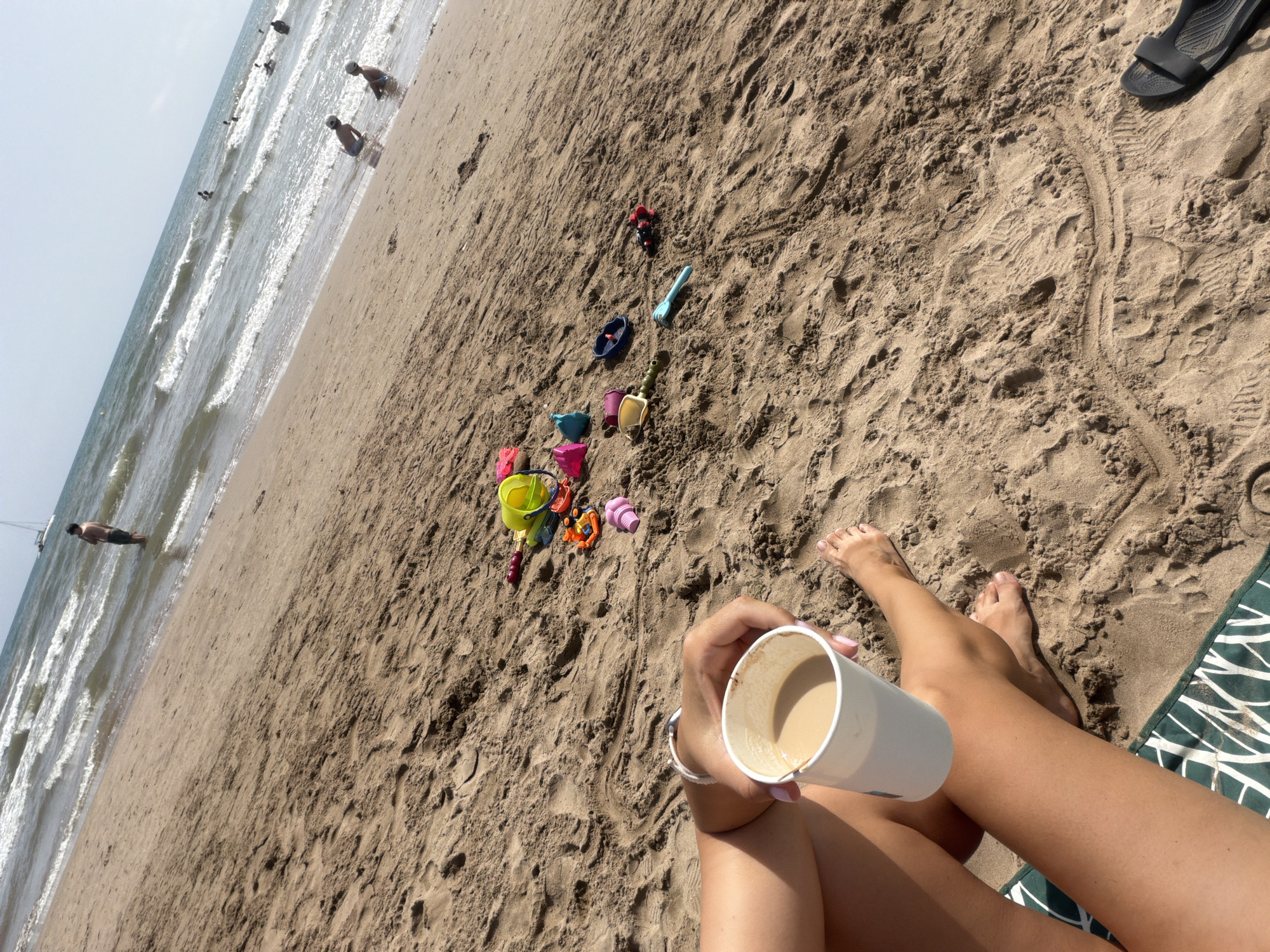 The image depicts a sandy beach scene with a body of water in the background. In the foreground, a person's legs and hands are visible. They are holding a white cup filled with a light-colored beverage, possibly coffee. Scattered on the sand are various children's toys, including buckets, shovels, and plastic toys. In the middle ground, several people are in the water, some appearing to be children sitting near the shore. Further back, a sailboat is visible on the horizon, and a person is standing in the shallow water. The overall environment suggests a sunny day at the beach.