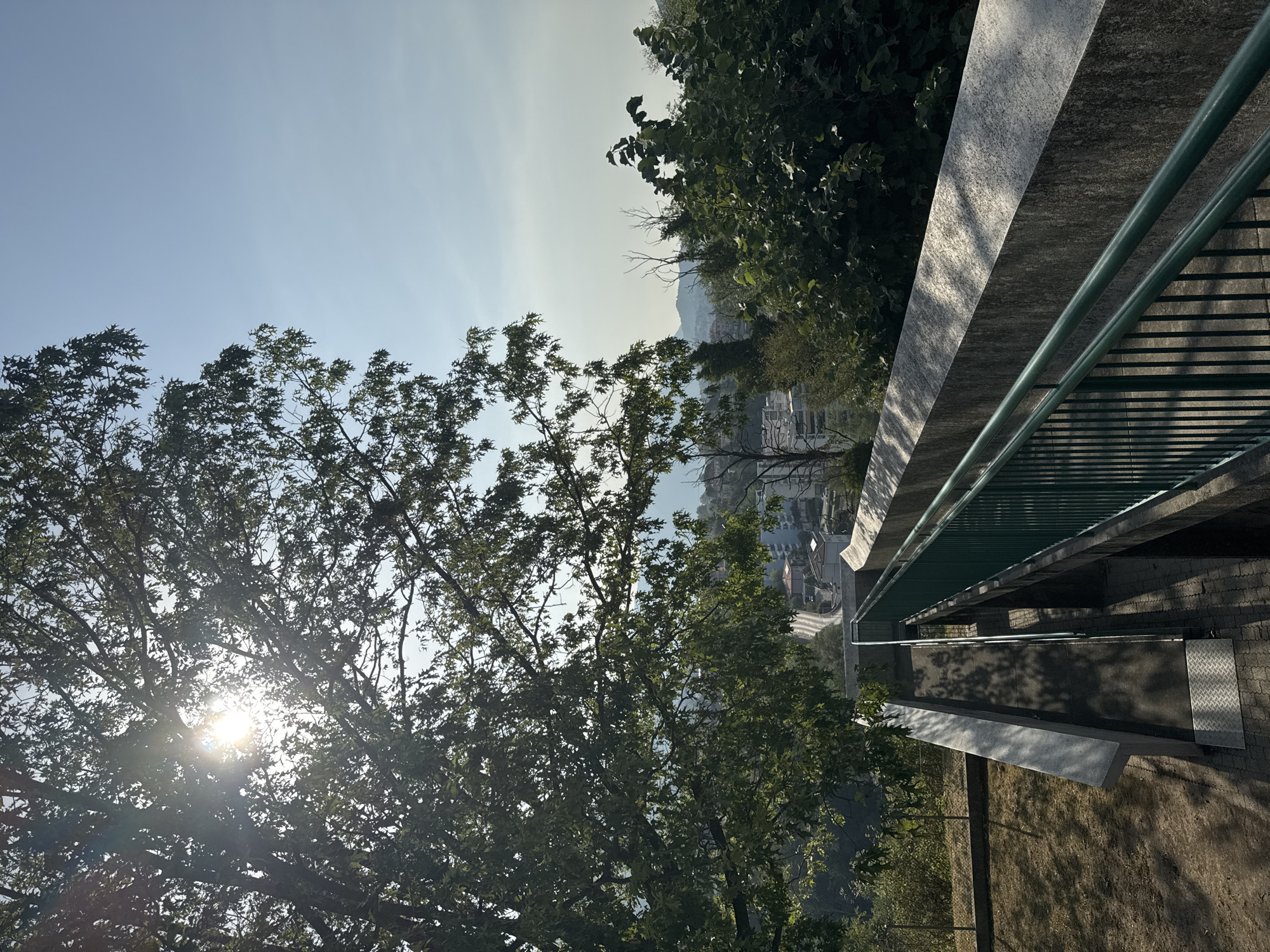 The image depicts an outdoor scene, likely a public walkway or staircase, with a view overlooking a town. In the foreground, a concrete structure with a green metal railing curves to the right, suggesting a pathway or bridge. The sun shines brightly through the leaves of a large tree on the left, casting dappled shadows on the ground. In the mid-ground, a town with buildings and a road is visible, nestled amongst greenery. The background shows hills or mountains under a clear blue sky. The lighting suggests it is daytime, possibly morning or afternoon, given the angle of the sun. No people are visible in the image. The environment appears to be an urban or semi-urban area with a significant amount of vegetation. The specific location in Balerna, Switzerland, is not explicitly indicated by any visual cues within the image itself.