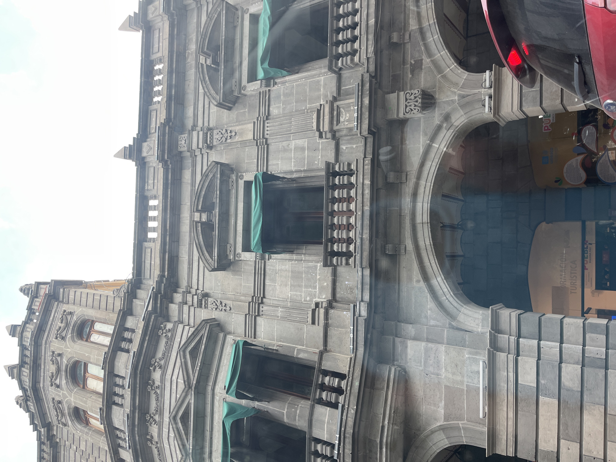 The image depicts a section of the Palacio Municipal de Puebla in Puebla, Mexico. The building is constructed of grey stone and exhibits ornate architectural details, including arched windows and decorative carvings. Some windows are draped with green fabric. A portion of a red vehicle is visible in the upper right corner of the image, obscuring parts of the structure. The photograph appears to have been taken from a low angle, emphasizing the height and grandeur of the building. A sign with "INFORMACIÓN TURÍSTICA" (Tourist Information) is slightly visible and reflected on a window.