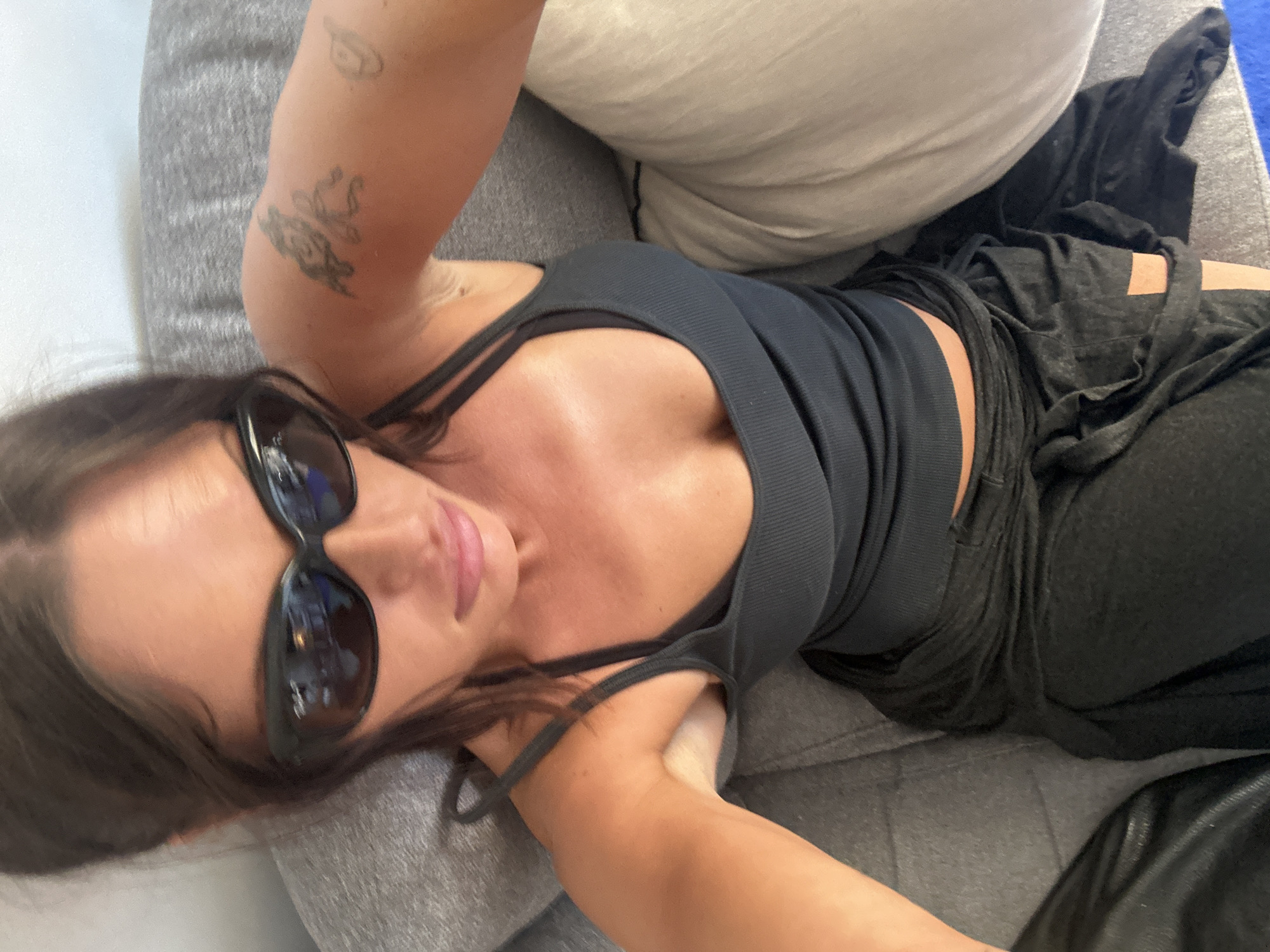 A woman is seated on a gray sofa, with a cream-colored pillow visible to her right. She is wearing dark sunglasses, a dark gray ribbed tank top, and dark pants. A tattoo is visible on her left upper arm. The setting appears to be indoors, though no specific location in London, United Kingdom can be determined from the visual information provided.
