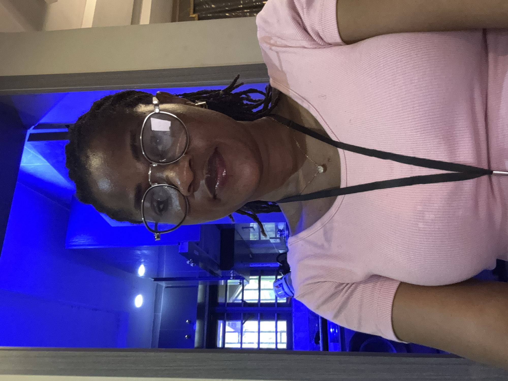 A woman with braided hair and glasses is looking at the camera. She is wearing a pink ribbed t-shirt and a necklace with a small pendant. A black lanyard is hanging around her neck. The background is dimly lit with bright blue lighting, suggesting an indoor setting, possibly a modern room or a nightclub. She appears to be indoors in Ajara, Nigeria.