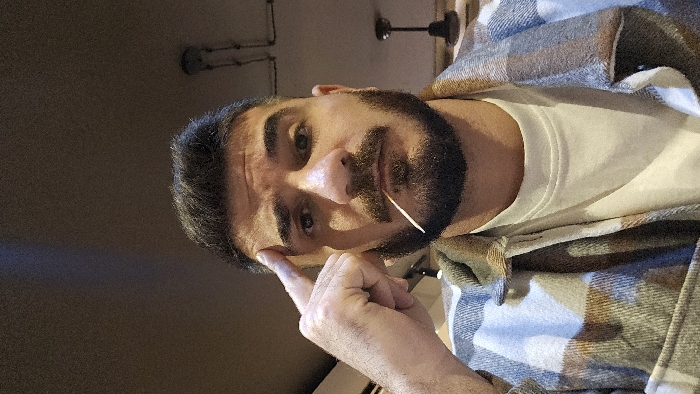 A selfie captures a man with dark hair and a beard, holding a toothpick in his mouth and pointing at his temple. He wears a white t-shirt under a plaid flannel shirt. The background is dark, with indistinct shapes. The image is slightly rotated.
