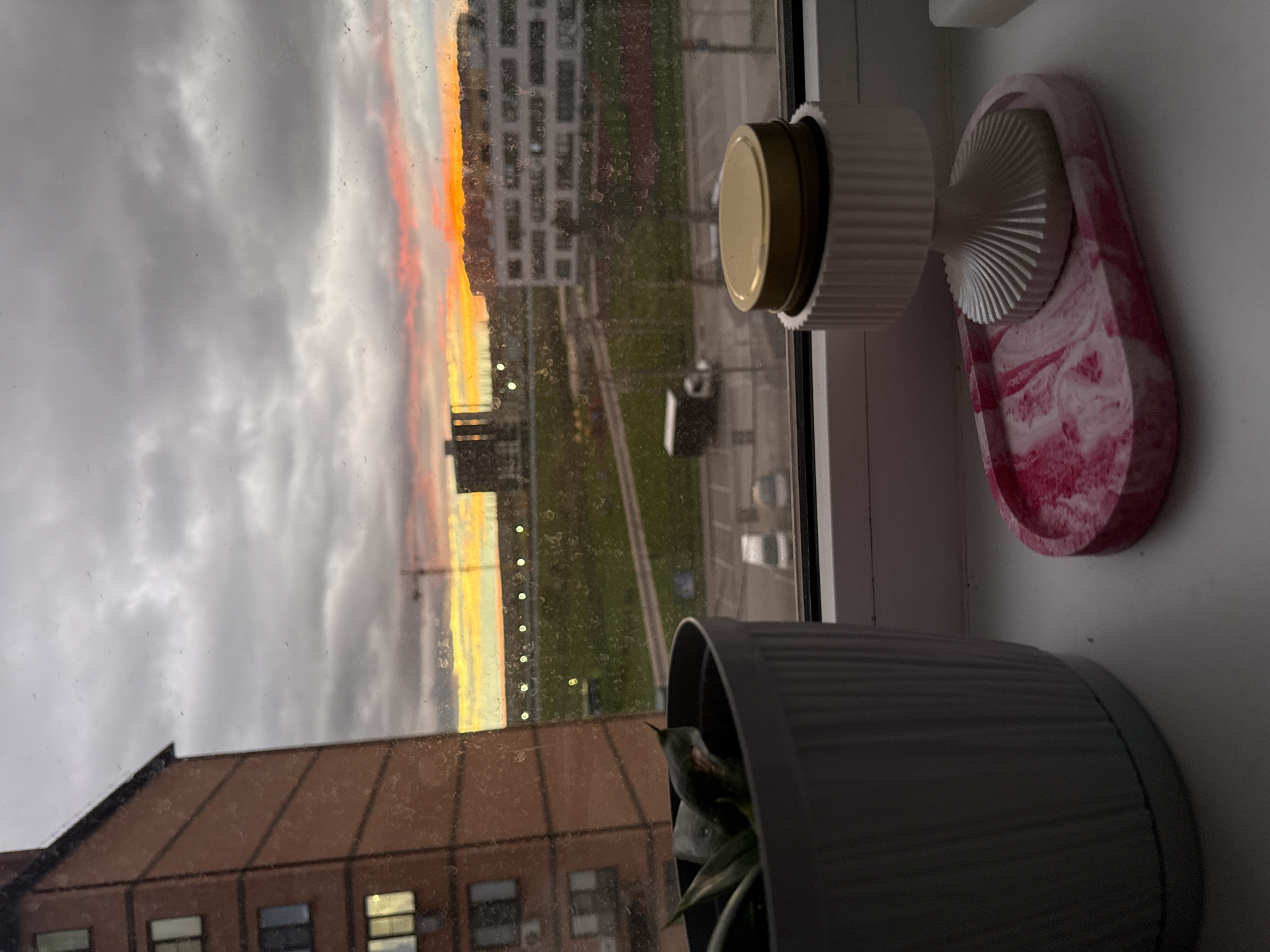 The image captures an indoor scene looking out of a window. Outside, a dramatic sky with grey clouds and a streak of orange and yellow sky, suggestive of sunrise or sunset, is visible. Buildings are seen through the glass, including an apartment block and a building with a pitched roof and multiple windows.
Inside, on the window sill, are a potted plant in a grey pot, a white dispenser or container with a gold lid, and a pink and white decorative tray with a white mushroom-shaped object on it. The scene appears to be in the city of Unknown, Unknown.