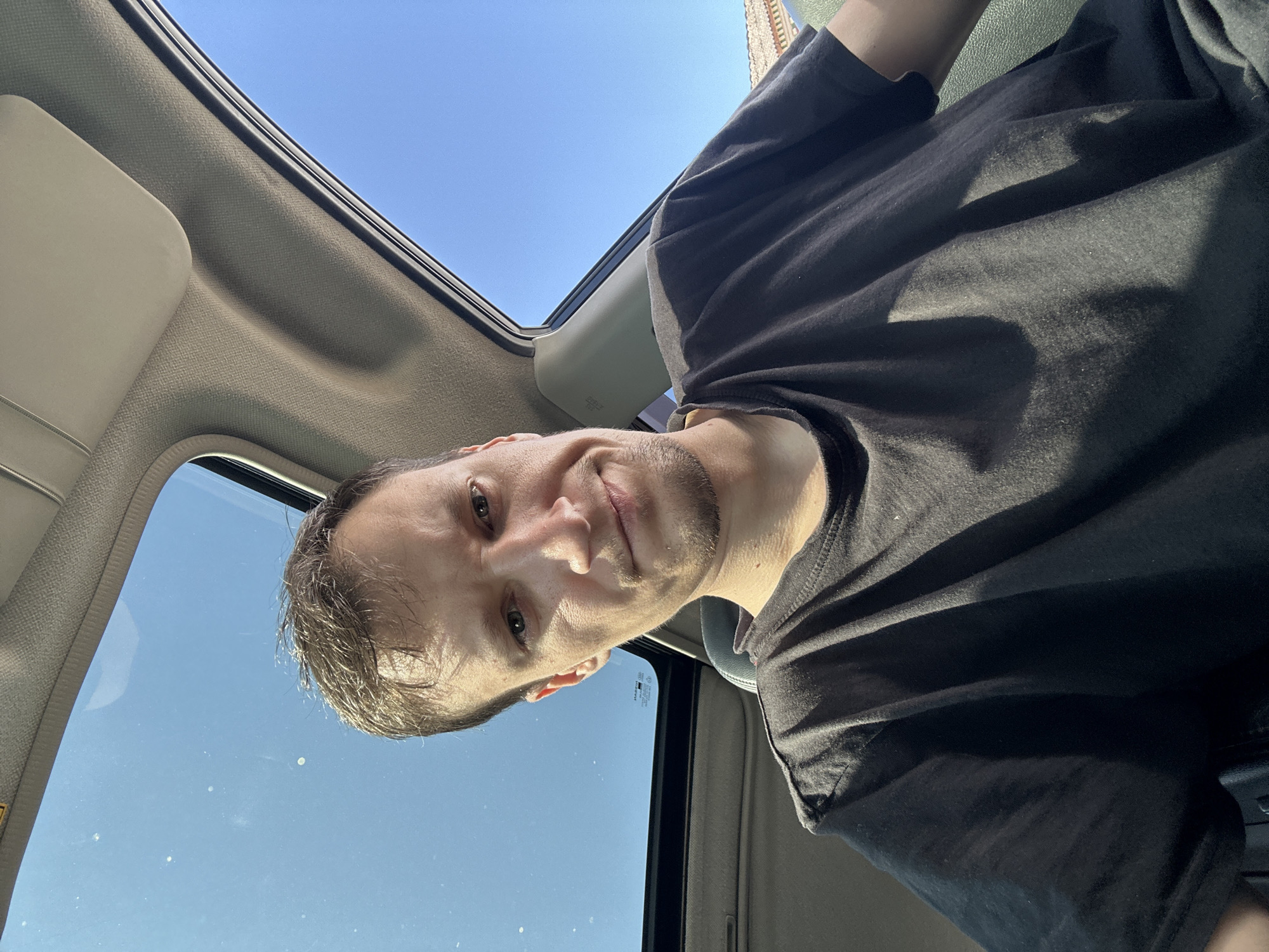 A man is in the front seat of a car, looking at the camera. He has short brown hair and is wearing a black t-shirt. The sunroof of the car is open, and the blue sky is visible. The interior of the car is visible, including the headliner and a sun visor. The scene appears to be in Shatava, Ukraine.