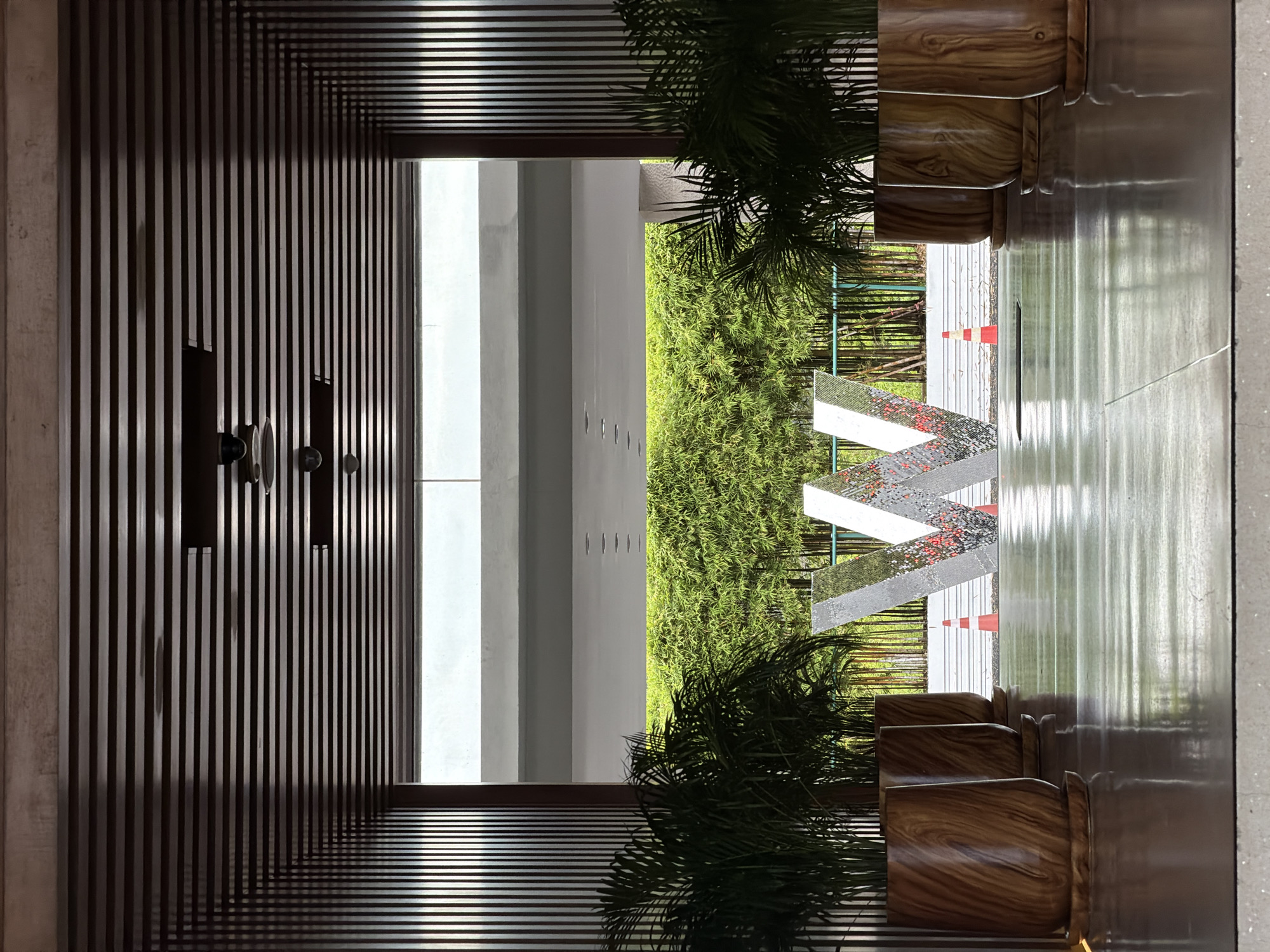 This image depicts the entrance area of a modern establishment, likely a hotel or resort, in Ko Samui District, Thailand. The perspective is from inside a covered walkway, looking outwards. The walkway has a dark, slatted wooden ceiling with recessed lighting fixtures. The walls of the walkway are lined with vertical wooden slats.
On either side of the walkway, large, wooden planters hold lush green tropical plants, possibly palms. In the center, beyond the walkway and in front of a dense bamboo-like foliage backdrop, stands a large, silver, three-dimensional letter "W". The letter is reflective, showing fragments of the surrounding environment. Two orange traffic cones are visible near the base of the "W".
The floor of the walkway is highly polished and reflective, mirroring the planters and the letter "W". The background reveals an outdoor area with the green foliage, a light-colored horizontal structure, and a bright sky, suggesting daylight. There are no people visible in the image. The overall impression is one of contemporary design and tropical ambiance.