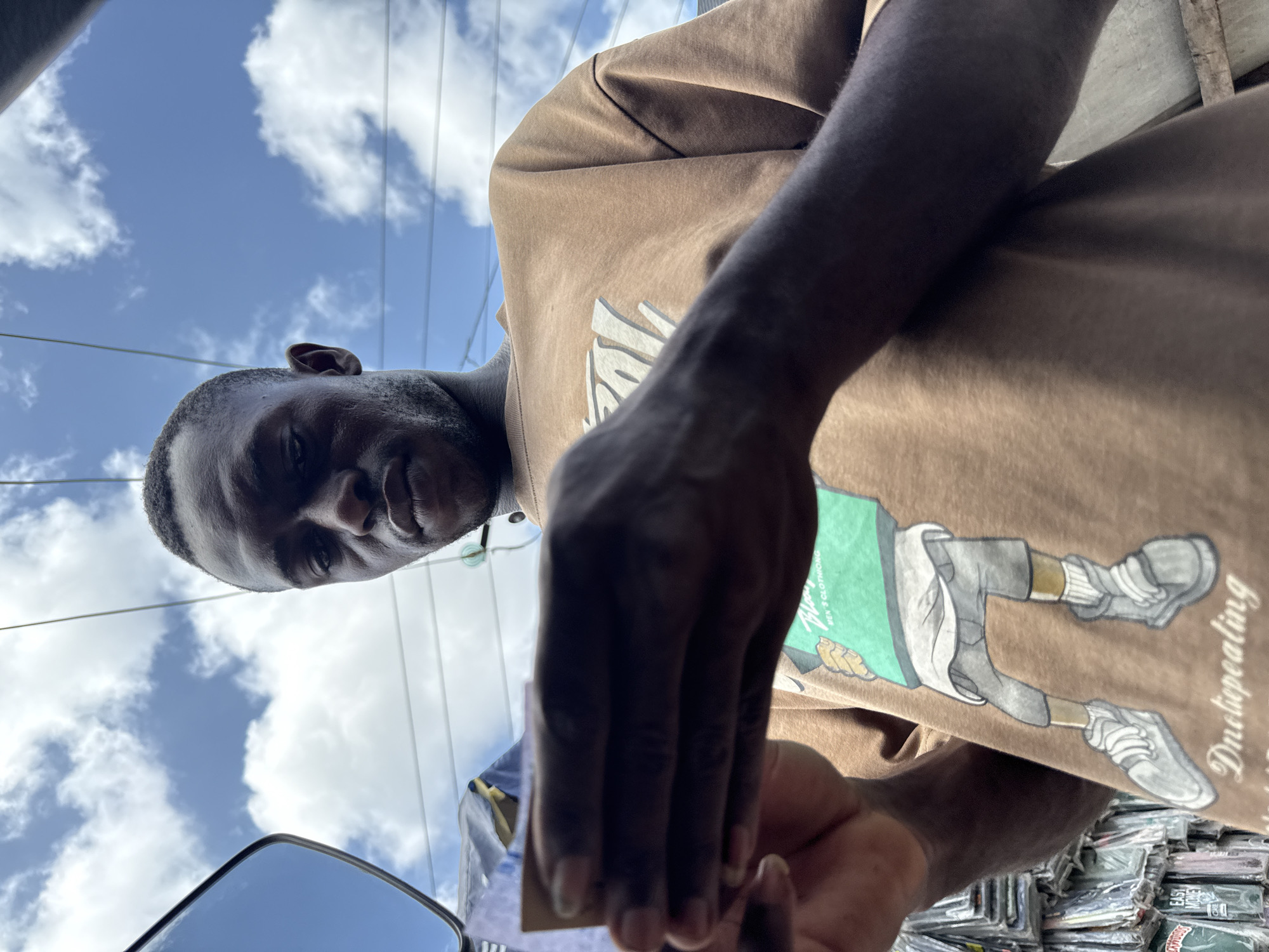 A man with dark skin is looking down at the camera from an upward angle. He has short hair and a beard. He is wearing a brown t-shirt with a graphic print of a cartoon character on it. The background shows a blue sky with white clouds and power lines. In the bottom left corner, there is a reflection of what appears to be a blue car in a mirror. In the bottom right corner, a pile of various packaged items can be seen. The scene is Ado, Nigeria, with the man holding what appears to be a small, rectangular object in his hands.