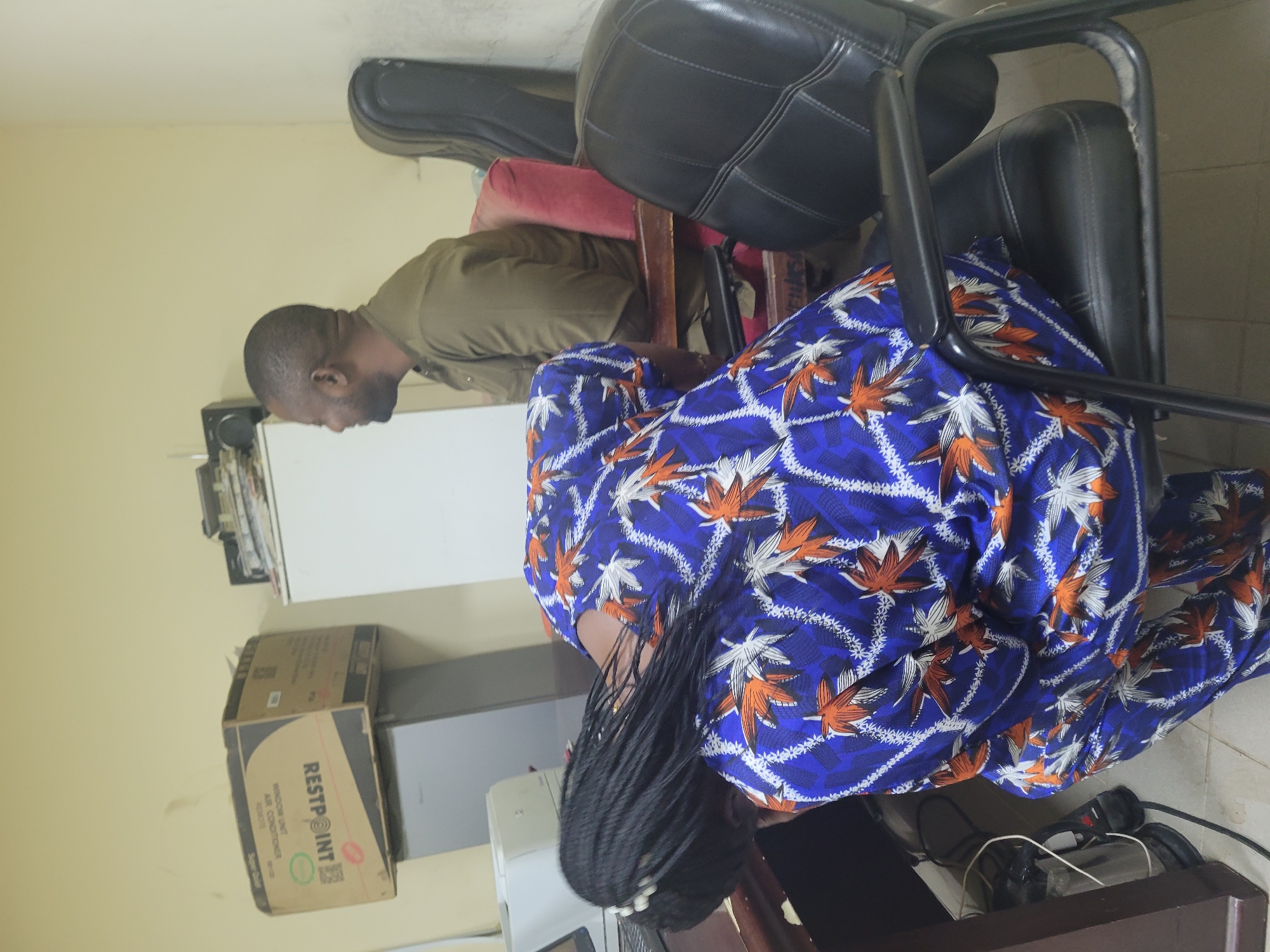 The image shows an indoor scene in what appears to be an office setting. A dark-skinned man with a shaved head, wearing a brown shirt, is seated in a chair. He is positioned behind a woman whose braids are visible. The woman is wearing a blue patterned outfit with white and orange leaf-like designs. They are both sitting in chairs with black cushions. A black guitar case rests behind the man. A "RESTPOINT" branded box, likely containing an air conditioner, is seen alongside a white surface or appliance. The background shows a cream-colored wall. The city and specific location are not definitively verifiable from the image.