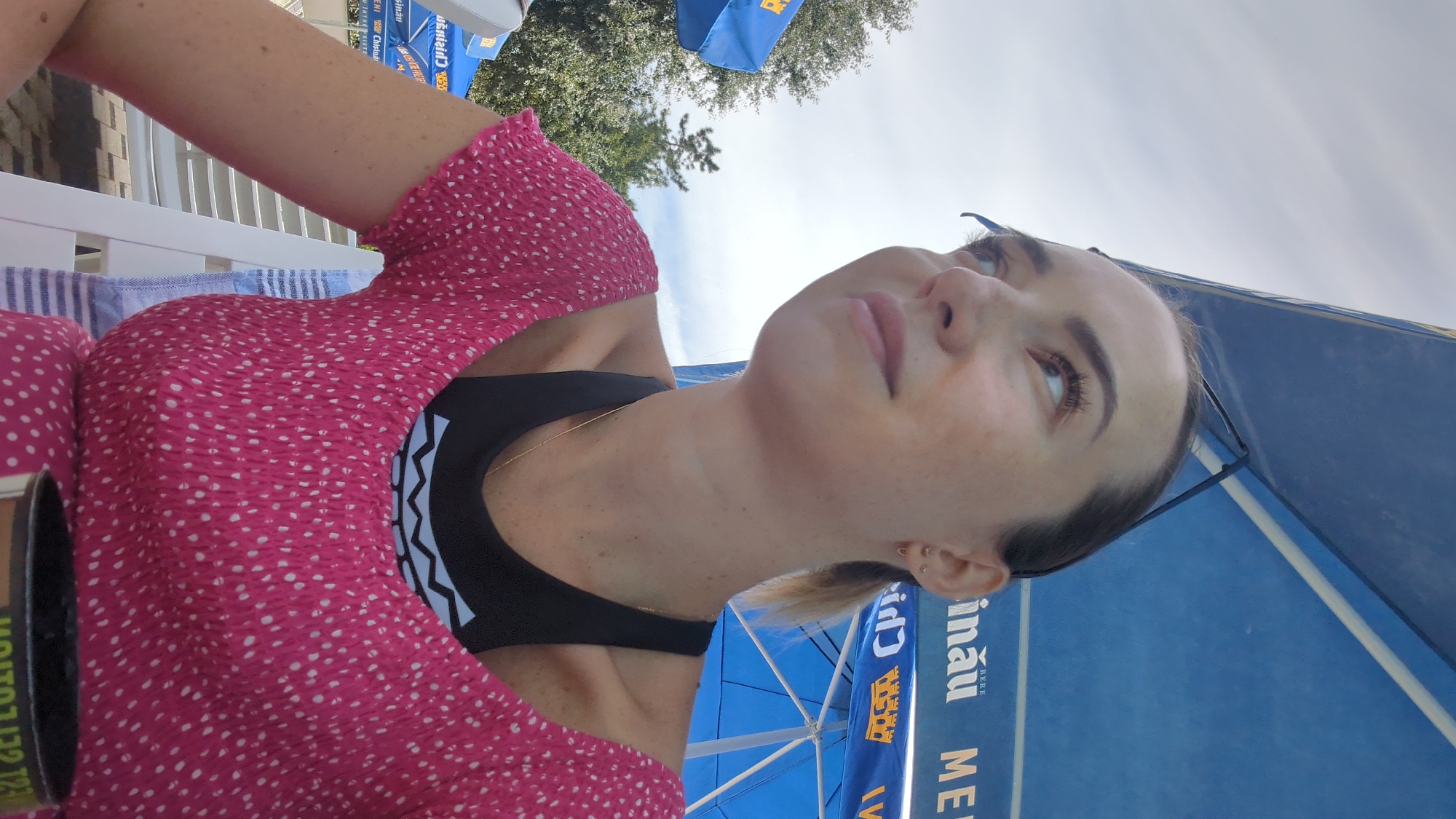 A young woman with light skin, dark hair pulled back, and freckles across her nose and cheeks is looking up and to her left, with her head tilted slightly. She is wearing a pink, polka-dot, smocked top over a black sports bra. A thin gold chain is visible around her neck. She has small earrings in her ears. The background is a bright, cloudy sky with a large blue umbrella overhead, which has text printed on it, and trees to the left. The woman appears to be seated on a white chair. The scene suggests an outdoor setting, likely a beach or pool area, given the umbrella and the woman's attire. The presence of the text on the umbrella suggests a commercial establishment. The setting appears to be in Vadul lui Voda, Moldova.