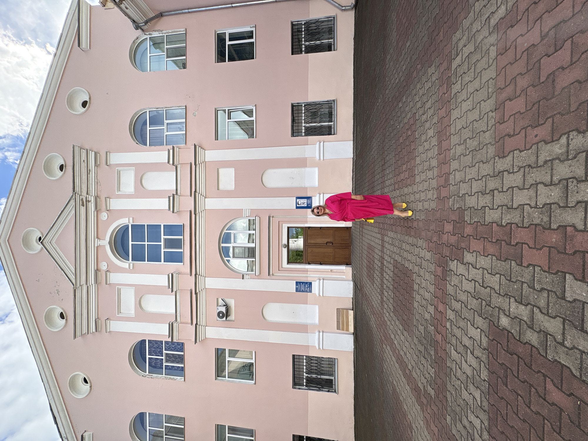 A woman in a bright pink robe stands on a paved walkway outside a pink building with arched windows. The woman is facing the camera with a slight smile. The building has multiple stories, with rows of windows and decorative architectural elements. The walkway is made of interlocking pavers in two shades of red and gray. In the background, the sky is visible with some clouds. The scene appears to be set during the daytime, with good weather. The image is taken from a slightly low angle, looking up at the building. There is a sign on the building above the woman's head with blue text and a small icon. The text on the sign is difficult to read. The overall impression is of a bright, cheerful scene.
