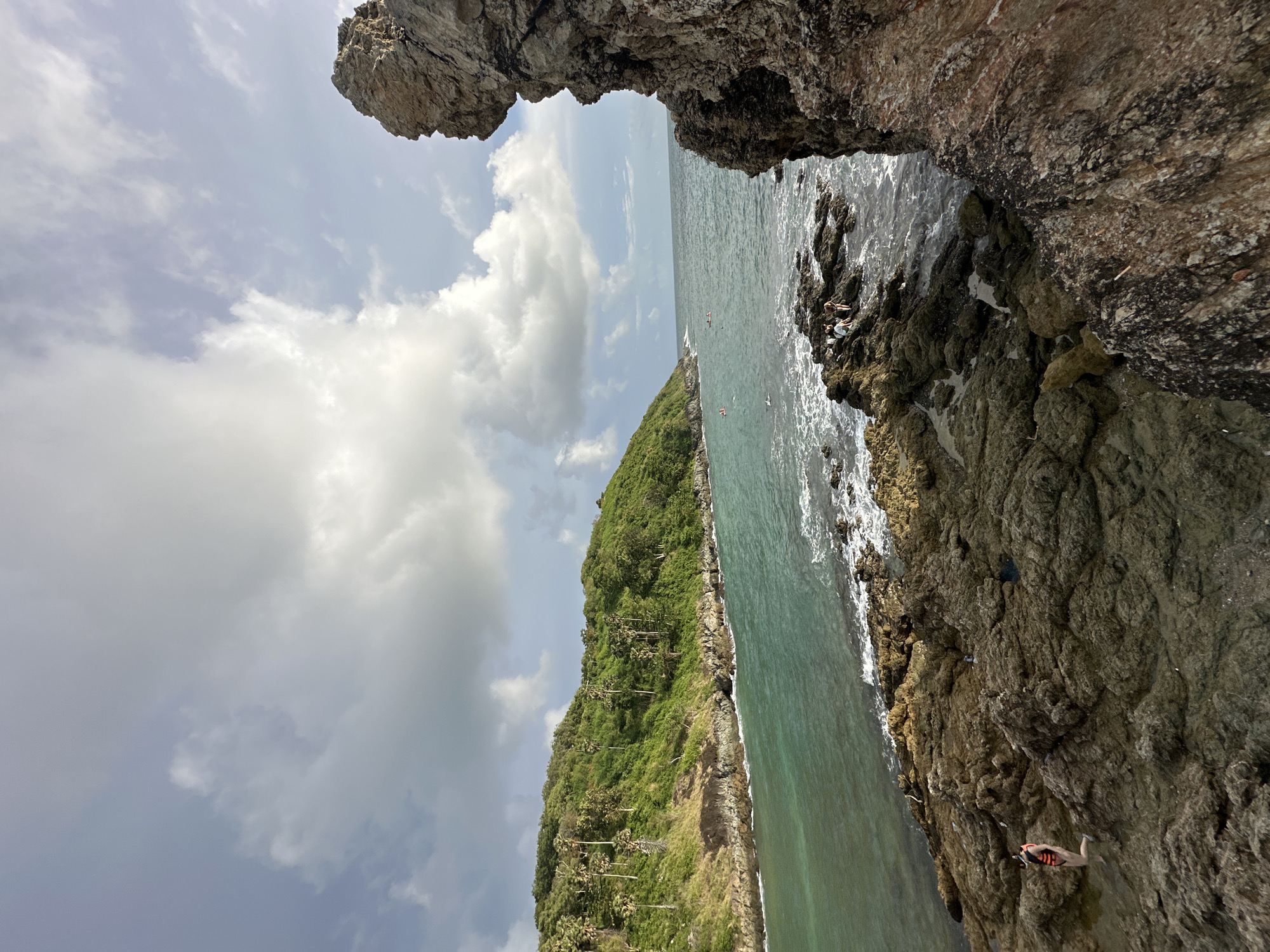 The image depicts a scenic coastal view of Rawai, Thailand, characterized by rugged rocky shorelines, lush green hills covered in palm trees, and the turquoise waters of the Andaman Sea. In the foreground, large, textured rocks dominate the right side of the frame, with smaller, jagged rocks forming the immediate shoreline. A person wearing an orange life vest is visible on the rocky shore in the lower left, interacting with the water. Further out in the sea, several individuals are seen swimming or kayaking, indicating recreational activity. The water is a vibrant teal, with white waves breaking against the rocks. The left side of the image features a sloping green hill densely populated with various trees, including tall palm trees. The sky is a mixture of light blue with scattered white clouds, suggesting fair weather with some cloud cover. The overall scene captures a natural, tropical landscape with human presence engaging with the environment.