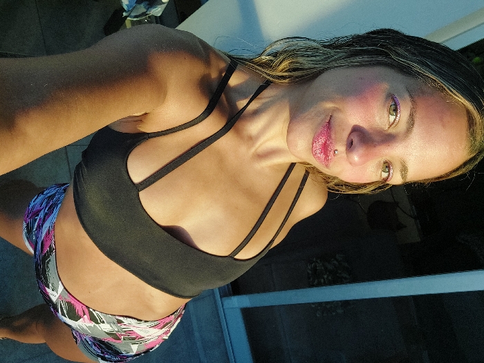 A woman poses in a black sports bra and patterned athletic shorts. The lighting is bright, casting shadows on her body. She has fair skin, light eyes, and blonde-streaked hair. Her lips are glossy, and a small stud is visible near her lip. The background is a mix of light and shadow, with hints of outdoor elements.