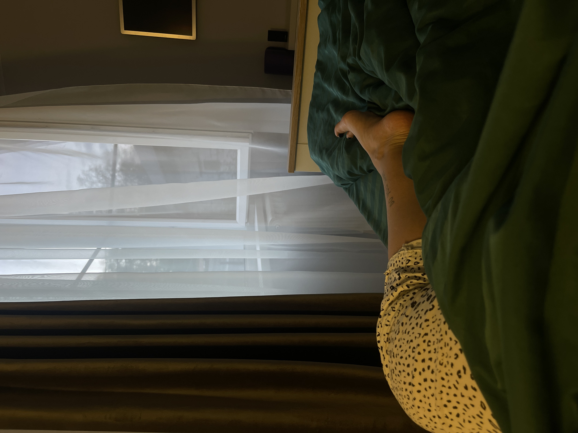 A view from within a room shows a leg extended from under a green comforter on a bed, indicating a person is lying in it. The person is wearing a tan and black patterned skirt. Beside the bed is a window covered with white sheer curtains, and behind that a darker brown curtain. The background includes a tan wall and an art fixture of a dark rectangle with a golden border. The image may have been taken in a room in Мосметаллоконструкция Онт, Russia.