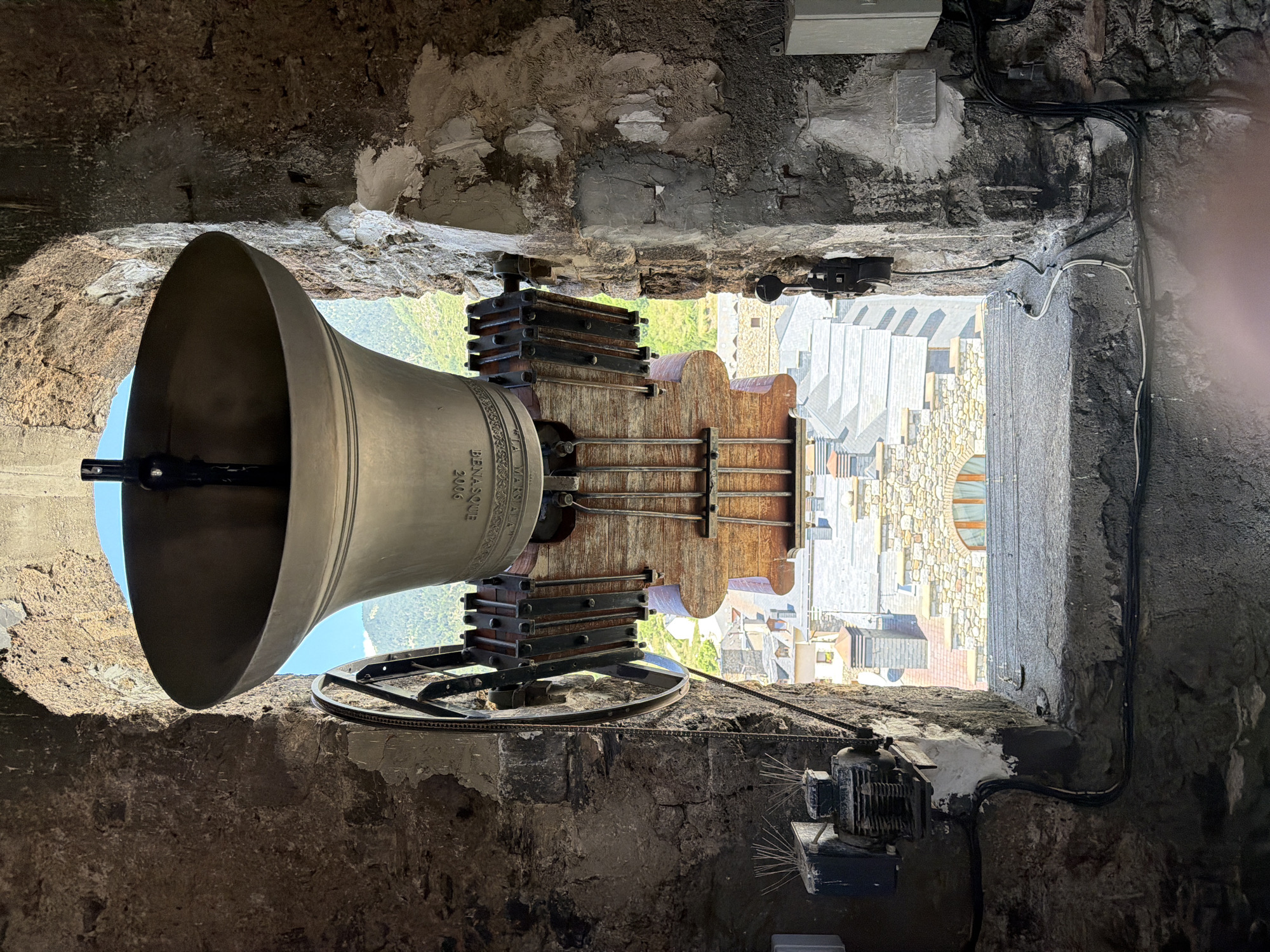 An extreme, low-angle, inverted shot looking up through a stone archway at a large bronze bell, suspended by metal and wood mechanisms. The bell is ornate, with text visible that reads "BENASQUE 2006". Below the bell, through a window opening, a town with stone buildings and gray roofs is visible, set against a backdrop of green mountains and a bright blue sky. On the left side of the archway, a motor and pulley system is attached to the stone wall, with wires running along the wall and down to the windowsill. On the right side, more wires are visible, along with a white electrical box. The stone walls of the archway are rough and weathered, with patches of cement and a few small spiky objects protruding from a box on the left. The image appears to be taken from inside a bell tower in Benasque, Spain, looking out and upwards.