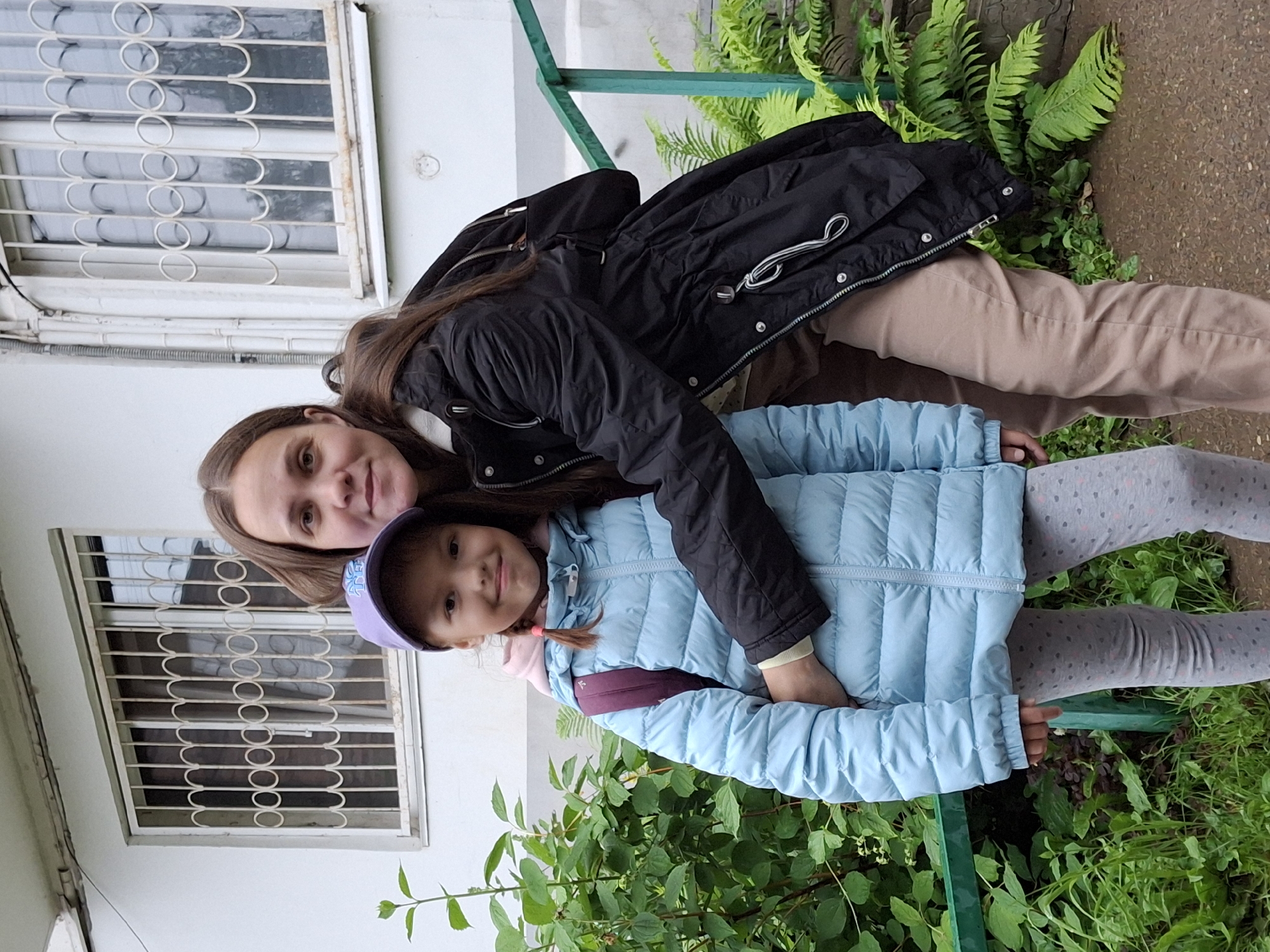 Two individuals, a woman and a young girl, are captured in a vertical, close-up photograph. The woman, with long brown hair, is positioned behind the girl, with her arm around the girl's shoulders. She is wearing a black jacket. The young girl, with dark hair and wearing a light blue puffy jacket and a purple cap, is in front of the woman. She is looking towards the camera with a slight smile.
They appear to be outdoors, with a white building with barred windows visible on the left side of the frame. Green foliage, including ferns and other plants, is present in the background, suggesting a natural or garden setting. A green railing or fence is also visible.
The lighting suggests it could be daytime, but the overcast sky or the shade from the building makes it difficult to determine the exact time of day. The presence of jackets indicates a cooler climate. The overall mood seems to be casual and affectionate, as the woman embraces the child. The location is identified as Chaikovsky, Russia. There is no visible text in the image.