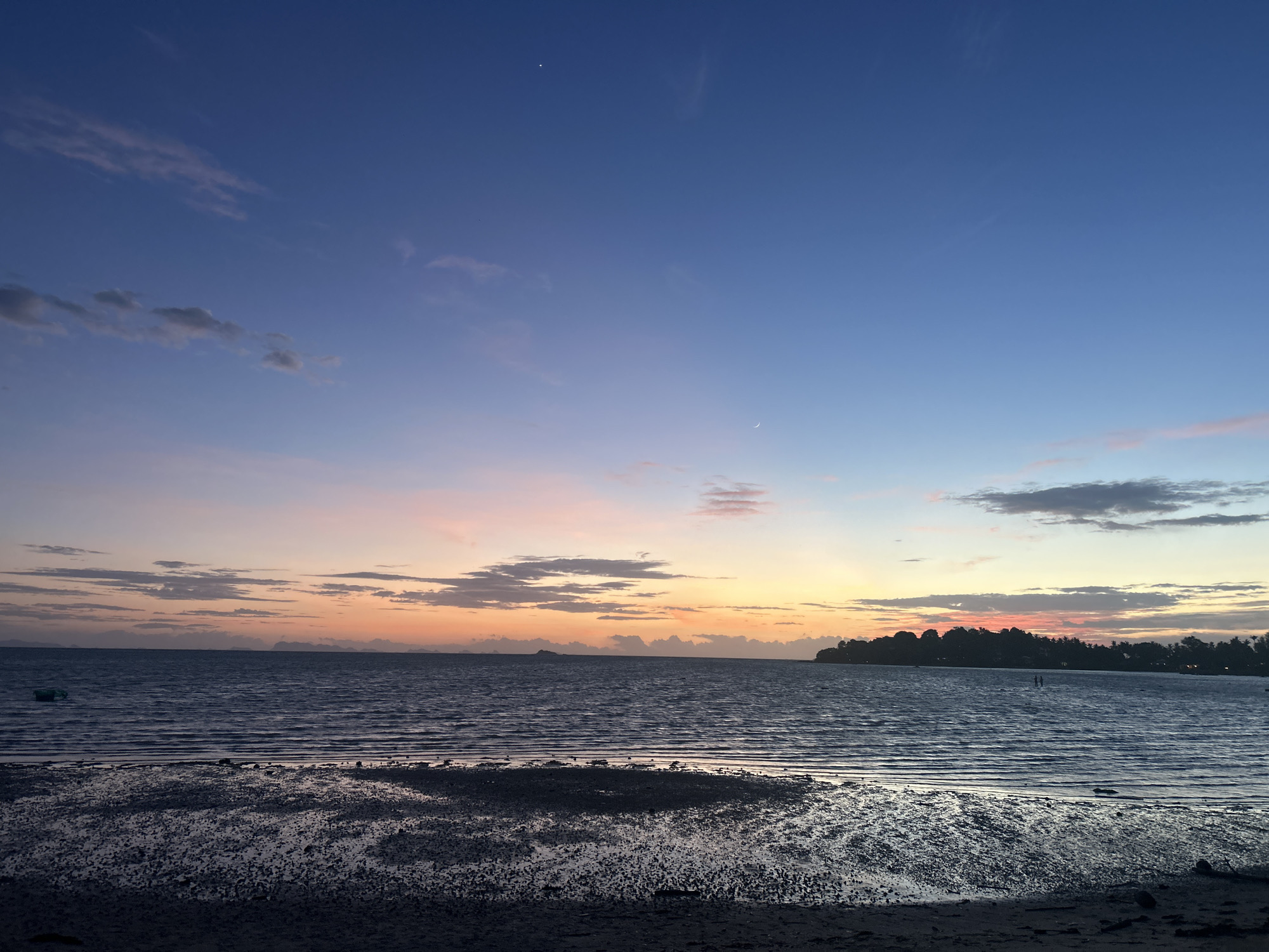A wide-angle, low-angle, eye-level shot captures a serene beach scene at dusk in Koh Phangan District, Thailand. The foreground is dominated by dark, wet sand with small rocks and ripples from receding waves, indicating a low tide. The middle ground features the calm, dark blue ocean stretching towards the horizon, reflecting the soft colors of the sunset. A small, green buoys is visible in the water to the left. In the distance, a silhouetted island covered with trees and vegetation forms the horizon. The sky transitions from a deep blue at the top to soft pinks and oranges near the horizon, painted with scattered, wispy clouds. Two faint celestial bodies, possibly stars or a thin crescent moon, are discernible in the upper portion of the sky. The overall atmosphere is tranquil and peaceful, characteristic of a sunset by the sea.