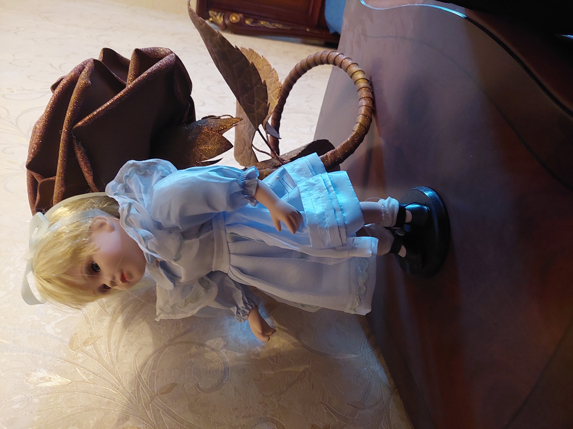 A doll with blonde hair and blue dress stands on a dark brown table. The doll is on a black circular base and has a white bow in its hair. Behind the doll is a large, dark red, glittery rose decoration attached to a metallic stem that curls into a circle. The background is a light-colored wall with a floral pattern. To the right of the doll, a portion of a wooden piece of furniture, possibly a headboard, is visible. The scene is indoors. The location of this image is not determinable from the visual information provided.