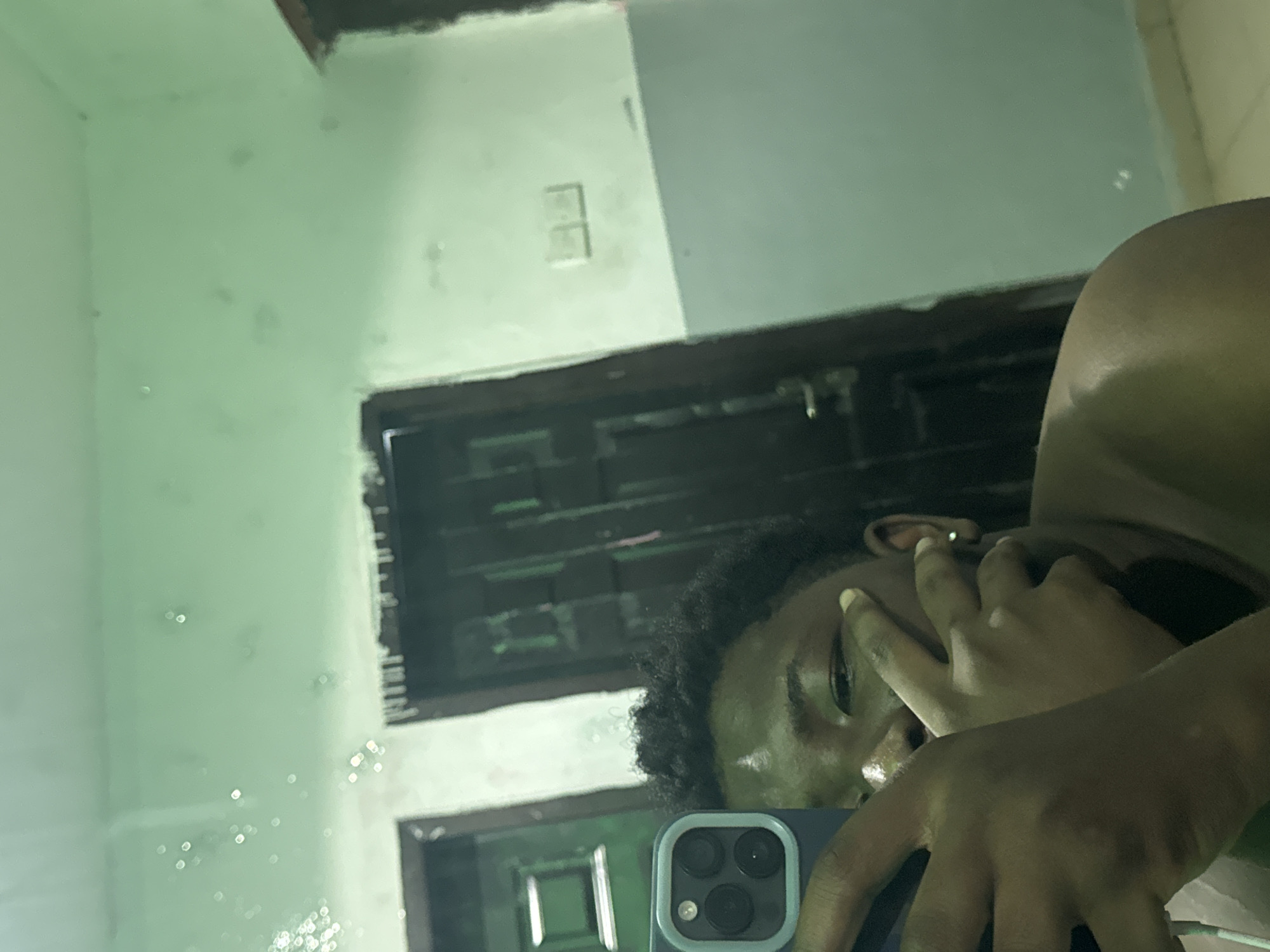 A person is taking a selfie in what appears to be a bathroom. The reflection shows a door, and possibly a light switch on the wall. The background color of the wall is a light green/mint shade. It's impossible to verify the location in Ipetumodu, Nigeria without additional information from the image.