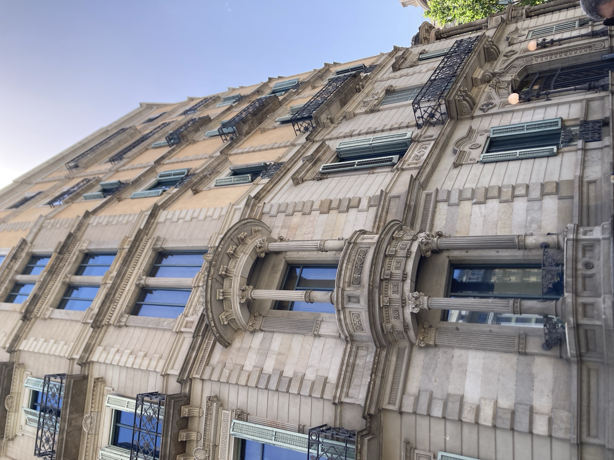 This media file is a low-angle, eye-level shot of the exterior of an ornate, historic building in Barcelona, Spain. The building features intricate architectural details, including decorative stonework, arched windows, and wrought-iron balconies. The facade is a pale, sandy color, with darker gray stone accents. Rows of windows are visible, some with open shutters, and others reflecting the blue sky. The building's design suggests an Art Nouveau or similar architectural style, common in Barcelona.
The perspective emphasizes the building's height and elaborate ornamentation. The lighting suggests it is daytime, with the sun casting shadows that highlight the building's textured surfaces. The sky is clear and bright blue, indicating fair weather. There are glimpses of green foliage at the top of the frame, hinting at trees or other greenery surrounding the building.
The scene is static, focusing solely on the architecture. No people are clearly visible, although a slight blur at the top right edge might suggest a person partially in frame. The overall impression is one of historical grandeur and detailed craftsmanship. The building appears to be a commercial or residential structure, likely in a well-preserved historical district of Barcelona. No visible text is present in the image.
