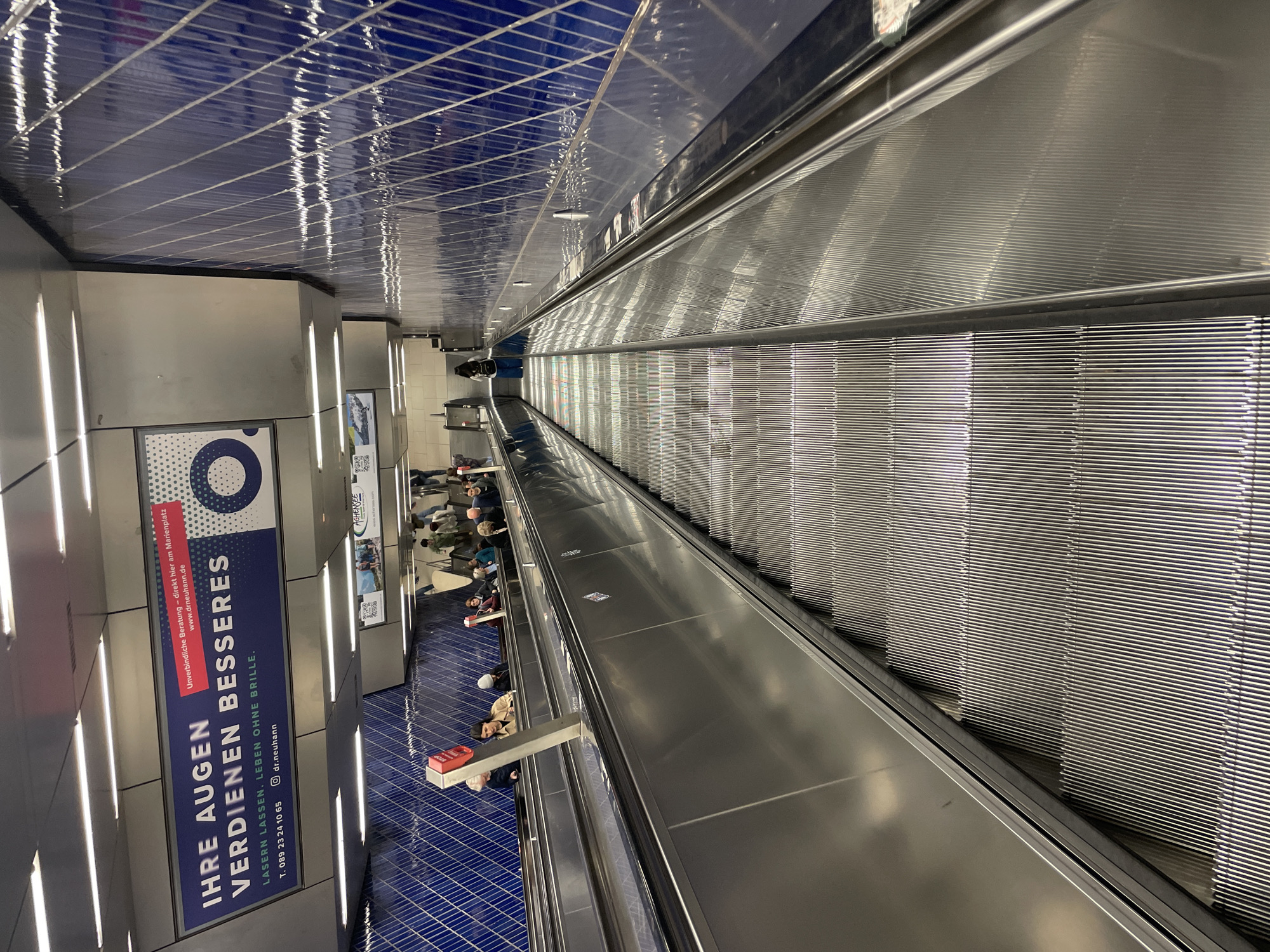 This image captures a wide, deep view of a modern underground station, likely a subway or metro, characterized by a long escalator and an adjacent moving walkway. The perspective is looking down the length of the passage.
The scene is dominated by striking architecture. Overhead, a glossy, dark blue tiled ceiling with a distinct grid pattern reflects the artificial lighting and elements below, creating shimmering streaks. The side walls are clad in metallic panels, featuring vertical strip lighting that illuminates the space. The floor on the left, where people walk, is also dark blue tiled.
On the right side of the image, a long, wide escalator descends, its metallic steps and side panels gleaming. To its left, a flat moving walkway also stretches into the distance, carrying passengers. Several people are visible on the left side, either standing on the moving walkway or walking on the adjacent tiled floor. They appear to be commuters or travelers, engaged in typical transit activities.
A prominent, illuminated advertisement is affixed to the metallic wall on the left. The text is in German and reads: "IHRE AUGEN VERDIENEN BESSERES" (Your eyes deserve better), "LASERN LASSEN. LEBEN OHNE BRILLE." (Have laser surgery. Life without glasses.). Below this, it offers "Unverbindliche Beratung - direkt hier am Marienplatz" (Non-binding consultation - directly here at Marienplatz) with contact details "www.drneuhann.de", "T. 089 23 24 10 65", and "@dr.neuhann". A smaller, partially obscured sign further down also shows "AGHENSEE".
The environment appears clean, well-lit, and spacious, suggesting an active urban transit hub. The time of day is indeterminate due to the artificial lighting.