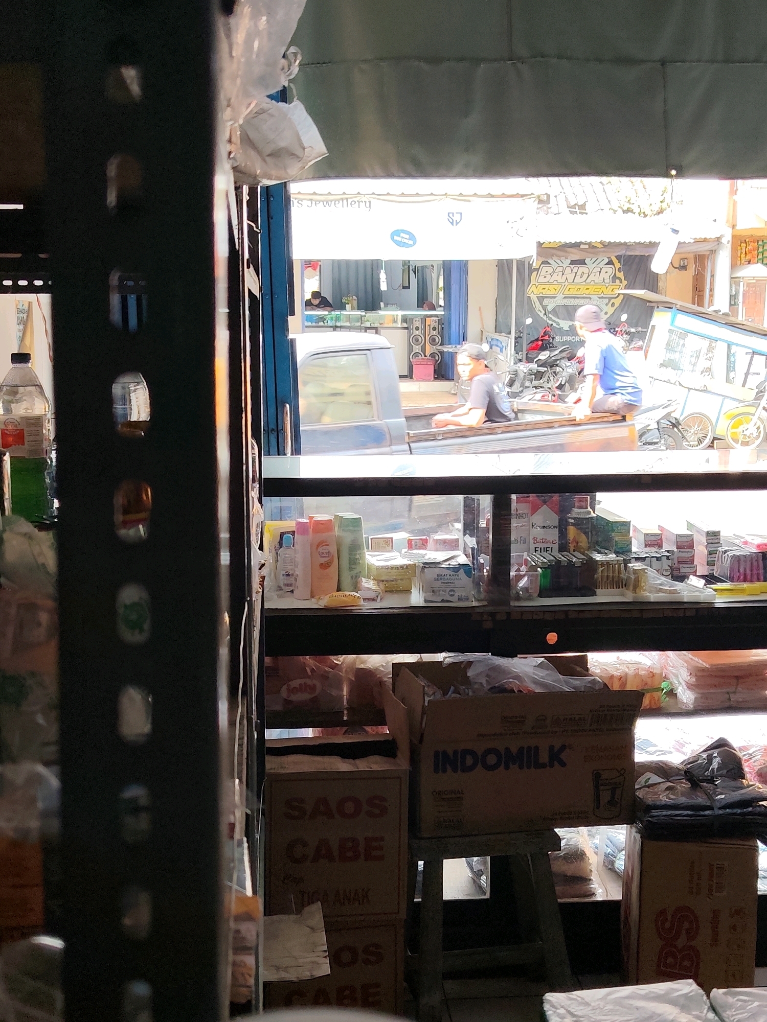 The image depicts a street-level view of a market or shop in Dampit, Indonesia. In the foreground, to the left, is a dark metal shelving unit with various items on display, including bottles and bags. To the right, through an open stall or shop front, a street scene is visible. A vehicle, possibly a pickup truck, is parked, with two individuals sitting in the truck bed. Behind them, motorcycles and a cart are parked. Further in the background, several shops line the street, with one displaying a banner that reads "BANDAR NASI GORENG". The shops appear to sell various goods, including what seems to be jewelry. The overall scene suggests a bustling commercial area on a sunny day.