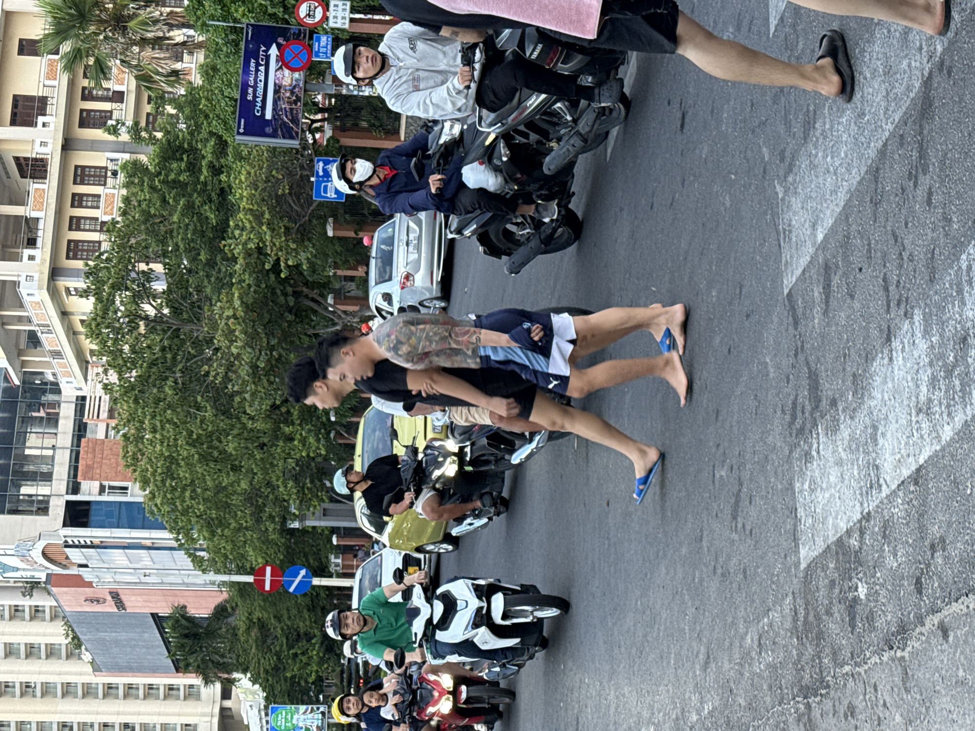 In Nha Trang Ward, Vietnam, a street scene unfolds with traffic and pedestrians. On the right side, two young men are walking across a crosswalk. One is shirtless and has extensive tattoos on his arms and torso, wearing blue and white patterned shorts and blue flip-flops. The other man, to his right, is wearing a light gray hoodie and dark shorts, with black slides on his feet. Behind them, two men on a black motorcycle are stopped at the intersection, one wearing a white helmet and mask, the other with a white helmet. To their left, a man in a green shirt is riding a white scooter, and a woman with a yellow helmet is on the back of a red scooter, accompanied by a child. Further in the background, several cars and motorcycles are visible on the street, and buildings of varying architectural styles line the background, interspersed with lush green trees. Traffic signs, including one with a red circle and a white horizontal bar, and another with a blue arrow, are present near the intersection. A billboard for "SUN GALLERY CHARMORA CITY" is also visible. The overall atmosphere suggests a bustling urban environment with active street life.