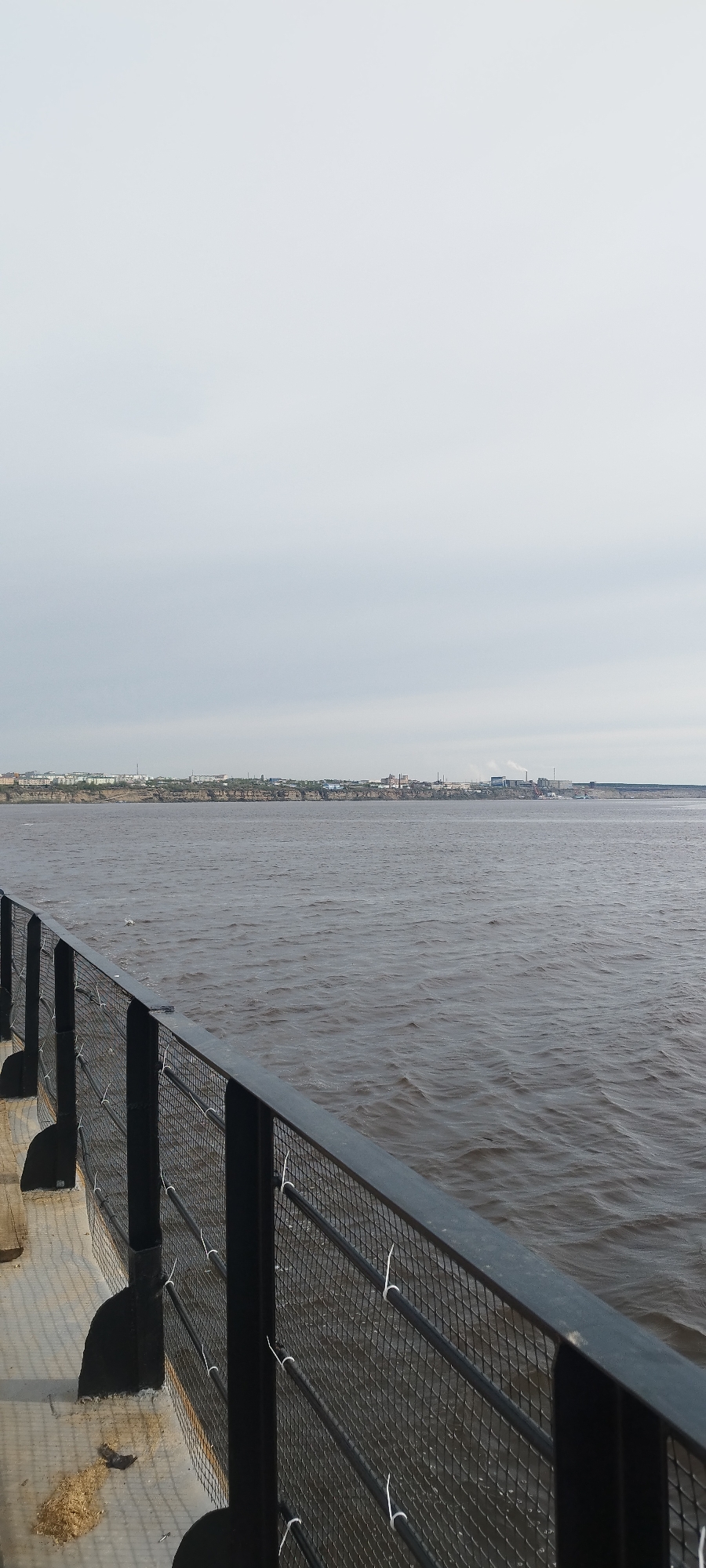 The image shows a view of a body of water, possibly a river, from behind a black metal fence. The water is dark and slightly choppy. On the opposite bank, a cityscape is visible with buildings and industrial structures, including what appear to be factories with smokestacks. The sky is overcast and gray. The fence is in the foreground and appears to be part of a walkway or deck. The white ties that are connected to the fence are holding something in place, but it is unidentifiable in the image. Given the description of Bestyakh, Russia, this is likely a view of the Lena River.