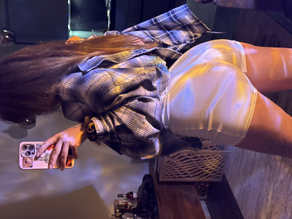 A woman wearing a plaid shirt and tight, shiny white shorts is taking a selfie. She's bending over, with her phone in her left hand, showcasing her backside. A white plastic basket is visible beneath her. A dark table with bottles is present in the background, and the image has a slightly dark and grainy quality.