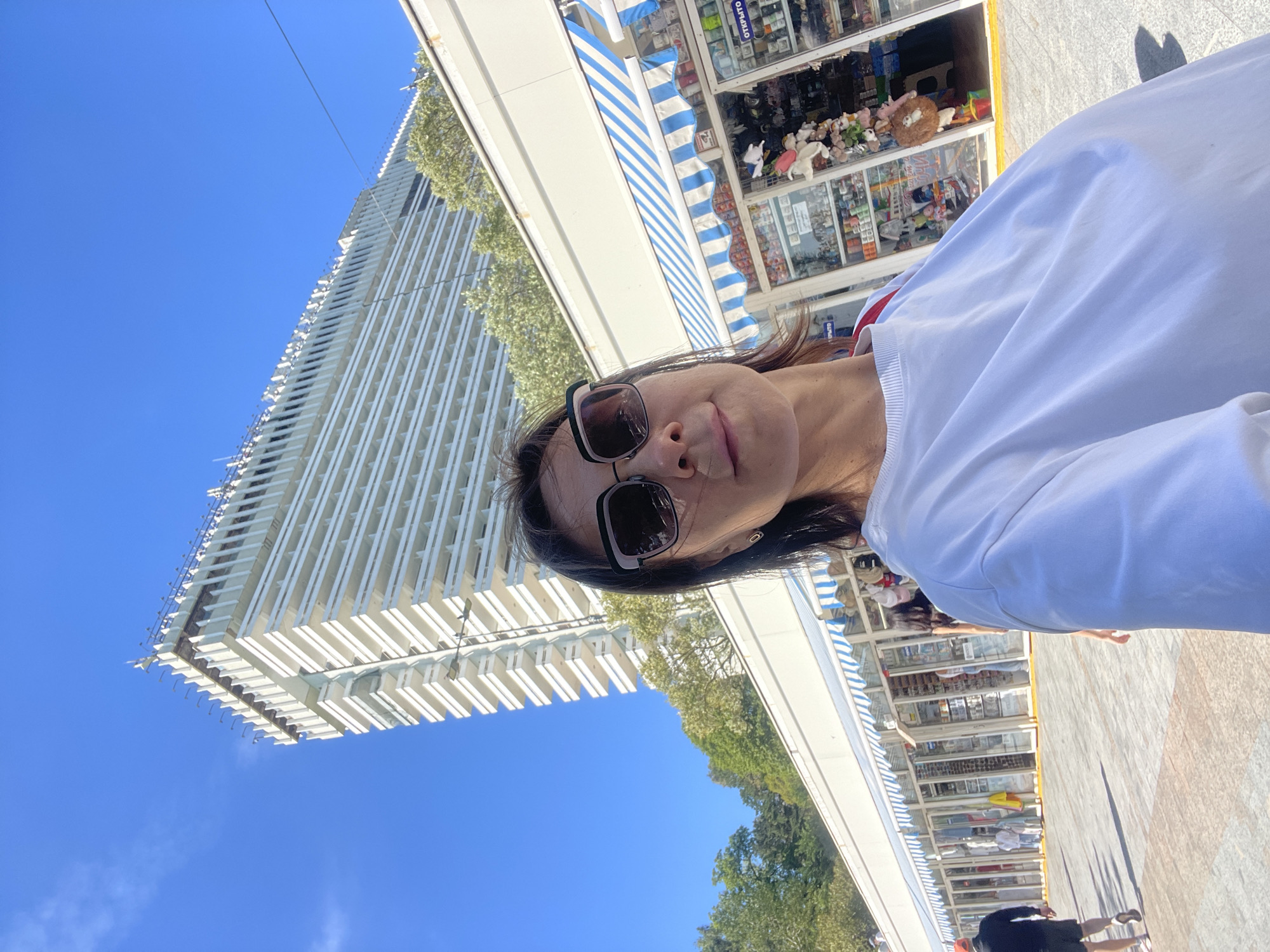 This bright, sunlit outdoor selfie captures a vibrant street scene, likely a promenade, in Sochi, Russia. The image is taken by a woman, positioned on the right side of the frame, looking towards the left with a slight smile. She has dark hair, wears dark-framed sunglasses, and a white long-sleeved top with a subtle red detail on the collar or shoulder.
Behind the woman, and stretching across the middle of the frame, is a row of small commercial establishments or kiosks. These shops feature distinctive blue and white striped awnings and glass display windows showcasing various goods, including an array of plush toys, small trinkets, and other merchandise. Visible on one shop sign is the Cyrillic word "ОТКРЫТО," meaning "OPEN." The foreground features paved ground, with the partial view of another person's legs and shadow in the bottom right, suggesting casual pedestrian activity.
Dominating the left side of the background is a tall, modern building with a striking architectural design, characterized by numerous white, vertical fins or balconies. Green foliage from trees is visible between the shops and the building. The sky above is a clear, brilliant blue, indicating bright, sunny weather. The strong shadows suggest it is daytime, likely mid-day or early afternoon. The overall scene depicts a pleasant, leisurely atmosphere in a bustling commercial and tourist area.