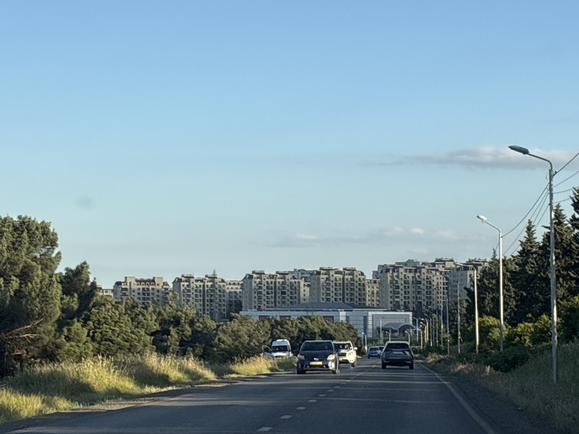 This media file depicts a view from a road leading towards a densely populated urban area with numerous apartment buildings. The buildings are tall and have a similar architectural style, suggesting a planned development. They are nestled behind a line of dense green trees, which are lush and appear to be in good health. The road itself is asphalt and has lane markings, indicating it's a significant thoroughfare. Several vehicles are driving on the road, including cars and a white van.
The time of day appears to be late afternoon or early evening, given the soft, warm lighting and the long shadows cast by the trees and streetlights. The sky is a clear, pale blue with a few wispy clouds, suggesting fair weather. The overall atmosphere is one of a developing urban landscape, with nature (the trees) existing alongside modern infrastructure. There are no people clearly visible in the foreground, but the presence of cars implies human activity. Streetlights are visible along the side of the road, spaced at intervals. The location is identified as Tbilisi, Georgia, which aligns with the architectural style of the apartment buildings and the type of vegetation commonly found in the region. No text is discernible in the image.