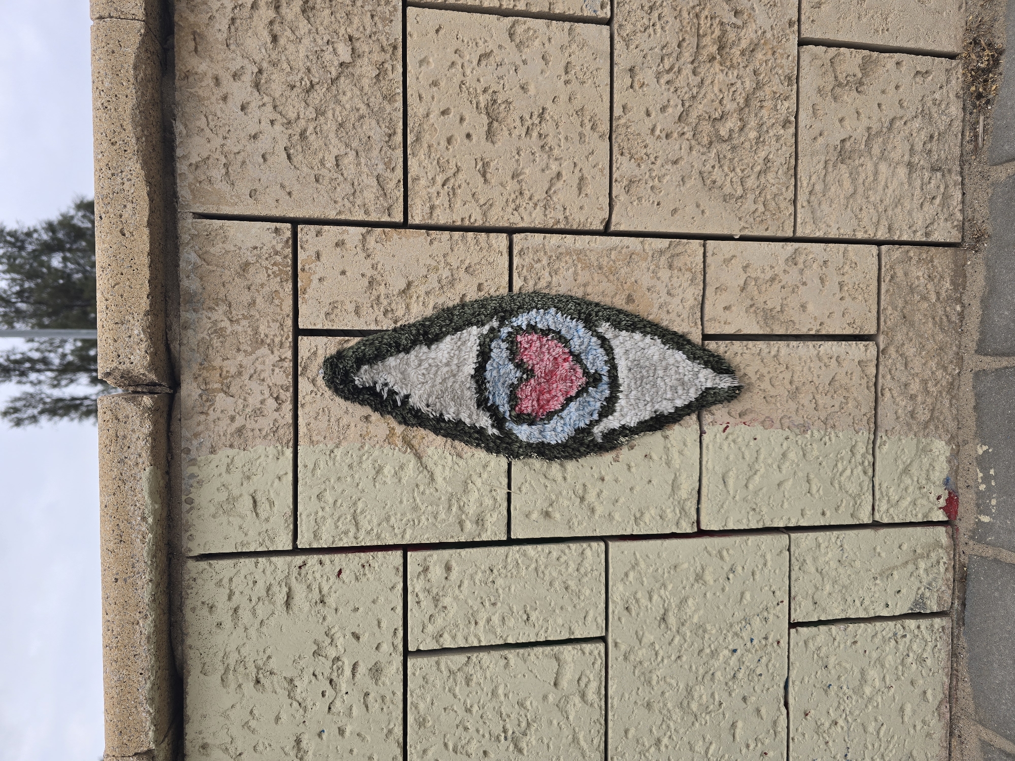 Here is a detailed description of the image:
The image shows a brick wall, with a piece of yarn bombing attached to it. The yarn bombing is in the shape of an eye with a heart in the center, and is affixed to a section of the wall that has been painted a light yellowish color. The rest of the wall is the color of natural stone. The wall appears to be part of a structure outdoors. Based on the style of the brickwork, the yarn bombing, and the general aesthetic, it is probable that the image was taken in the city of Tel-Aviv, Israel.