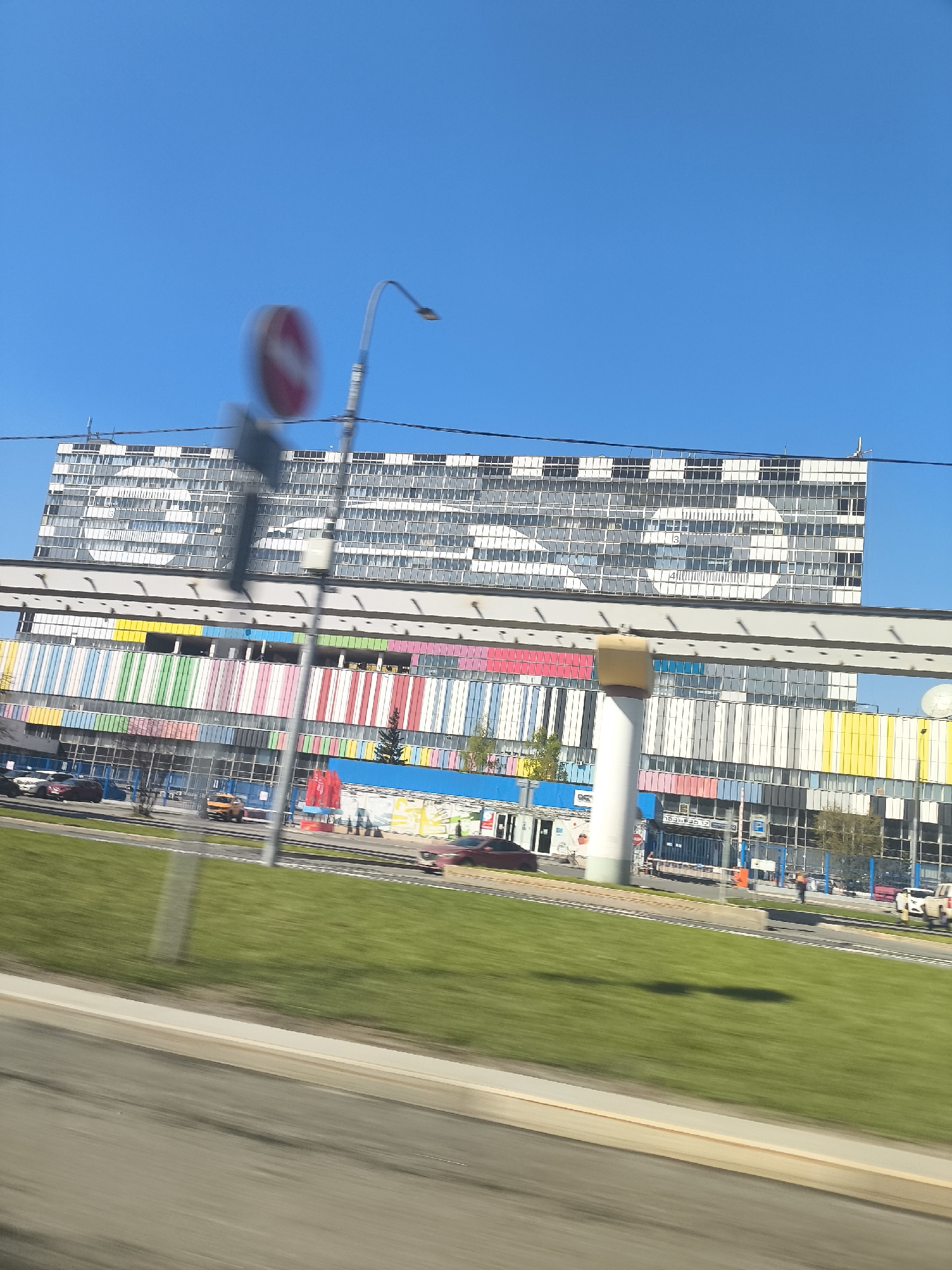The image shows a view from a moving vehicle of a large building complex in Khimki, Russia, under a clear blue sky. The top building is tall and rectangular, featuring a grid of windows that collectively form the letters "VTB." Below it, there is a colorful building with vertical stripes in various colors, with a monorail track running along its top. A street runs in front of the buildings, with sparse traffic consisting of cars. A grassy area is visible between the street and the vantage point of the image. A traffic sign and streetlight are blurry in the foreground, indicating the motion of the vehicle from which the photo was taken.