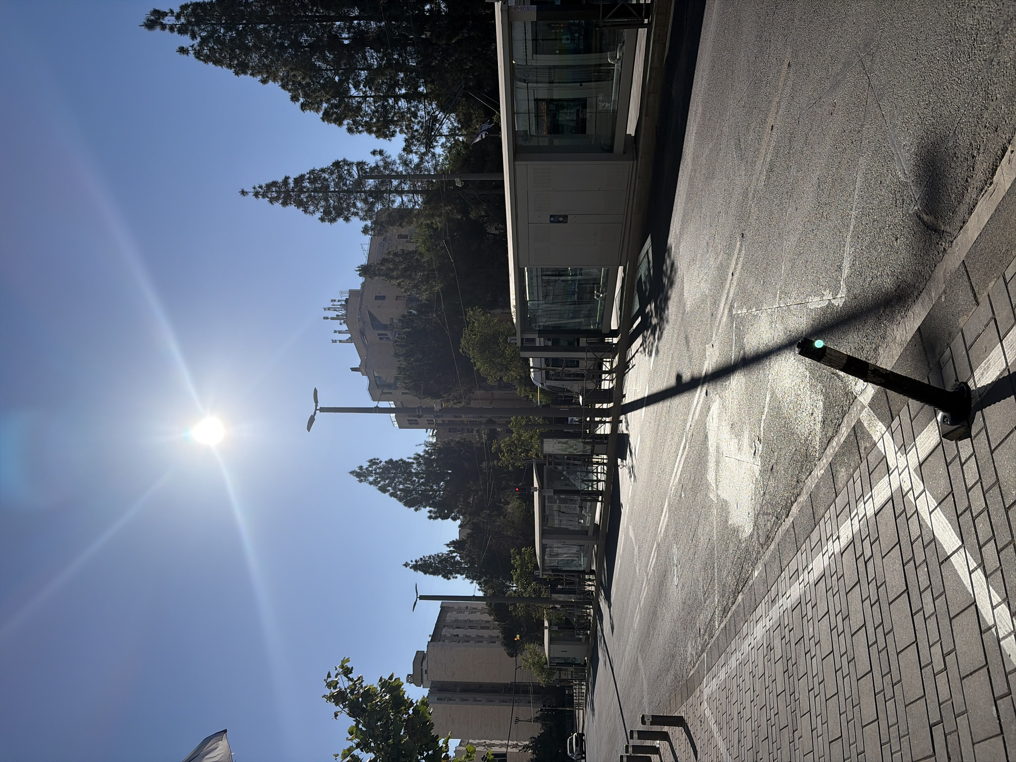 The image shows a street scene in Jerusalem, Israel on a sunny day. The perspective is from the side of the road, looking towards a row of modern bus stops. Buildings, including apartments and institutional structures, are visible in the background among trees. The sun is bright in the sky, creating lens flare and long shadows. The sidewalk is paved with grey brick and bordered by a white line. A black bollard with a green light is in the foreground, casting a sharp shadow across the road. The road surface is asphalt with some surface wear. No people are visible.