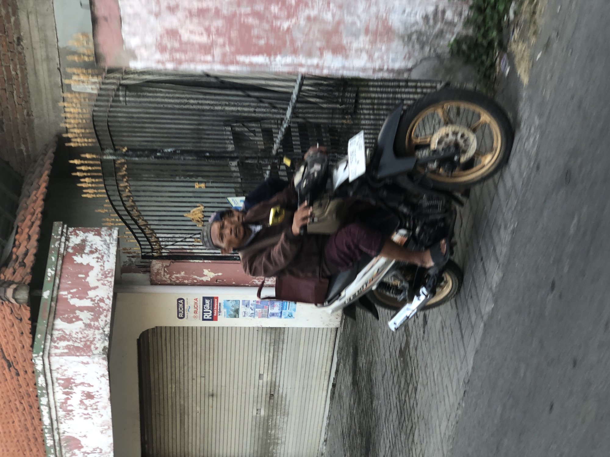 A man rides a motorcycle down a street in Sentul, Indonesia. The man is wearing a brown jacket, a maroon sarong, and a black skullcap. He has a bag on his shoulder. The motorcycle is black and silver. The street is paved with bricks. In the background, there is a building with a corrugated metal garage door and a gate. The building is painted red and white, and the paint is peeling. There are advertisements on the wall next to the garage door. The sky is overcast.