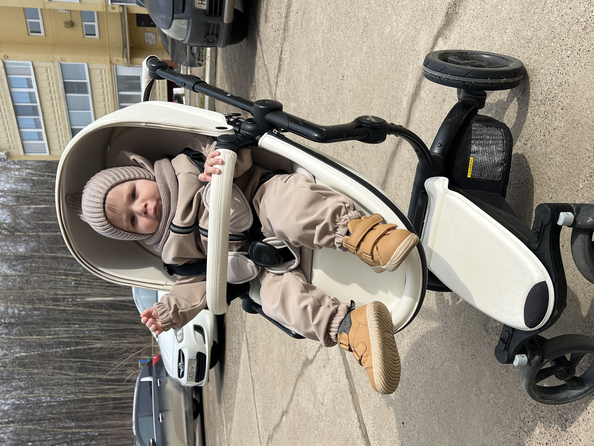A young child sits in a modern white stroller on a paved surface, seemingly outdoors. The child is wearing a beige outfit, including a knit hat, jacket, pants, and brown shoes. The stroller has a black frame and wheels. In the background, a building with yellow walls and multiple windows is visible, along with parked cars. The license plate of a nearby vehicle, "B820TM159", is visible, pinpointing the location as Noviy, Russia. Bare trees are also seen in the distance, suggesting a late autumn or early spring day.