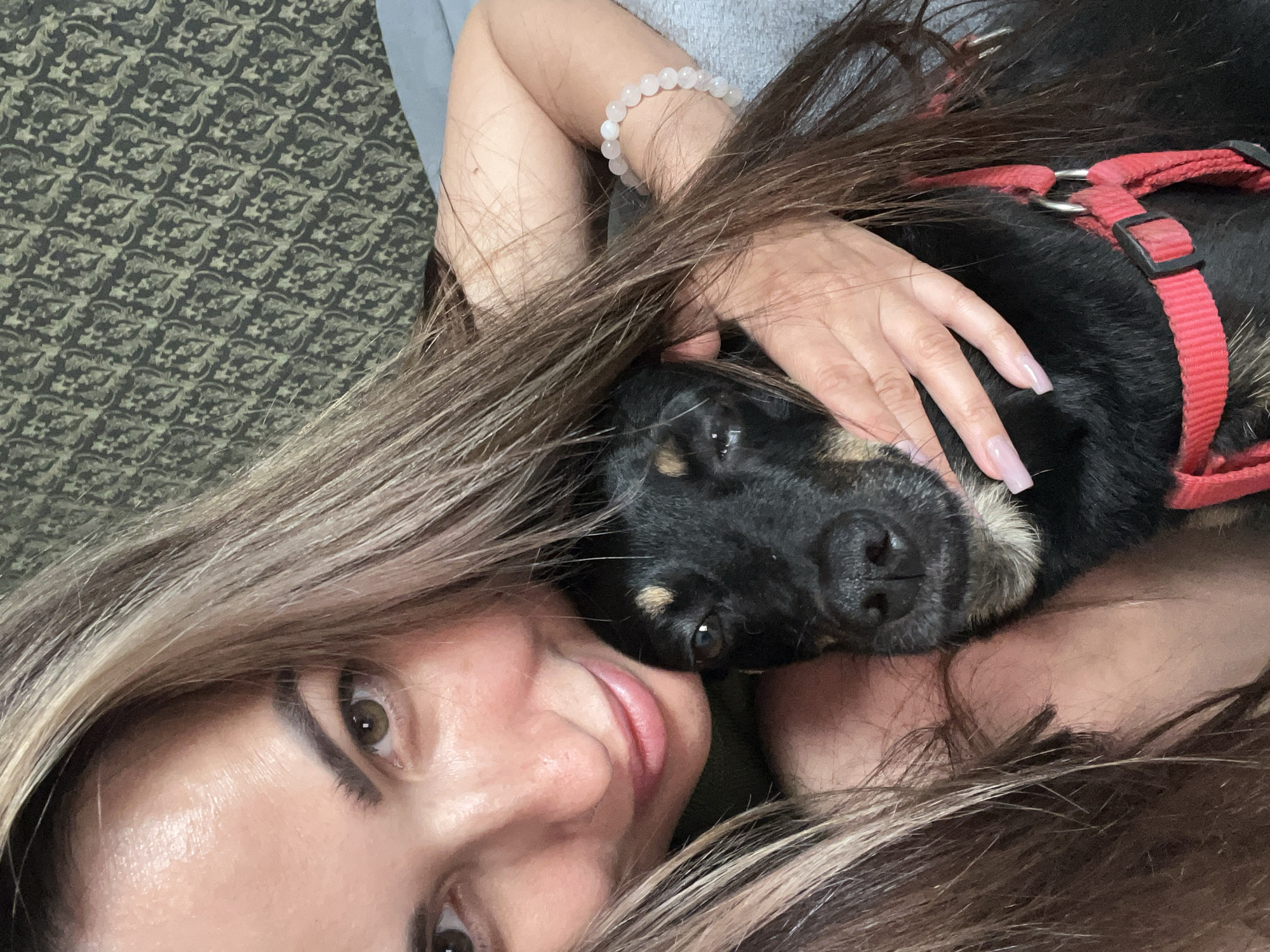 In a close-up selfie, a woman with long, light brown hair is embracing a black dog. The woman's face is turned towards the camera, with her eyes looking forward. She is smiling slightly, with her lips parted. Her left arm is wrapped around the dog, and her right arm is resting on its head. The dog's head is tilted up, and its eyes are looking forward. The woman is wearing a green top and a bracelet on her right wrist. The dog is wearing a red collar. The background is a patterned wall. The lighting is soft and diffused.