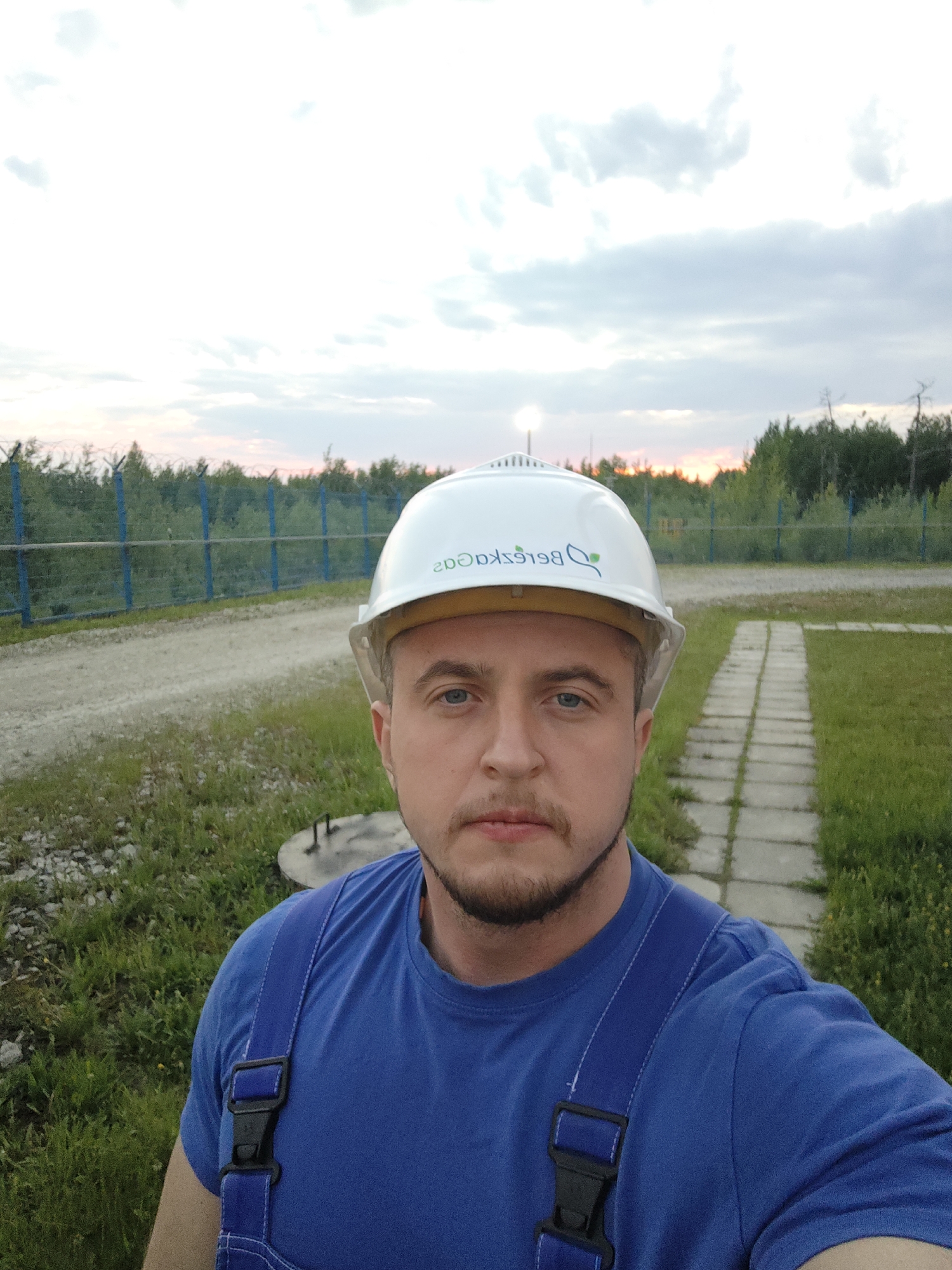 This image captures a close-up, selfie-style portrait of an adult male outdoors, presumably at an industrial or utility site in the city of Unknown, Unknown. The man, with fair skin, light blue eyes, and a short, dark beard, is looking directly forward. He wears a white hard hat with a mirrored logo featuring a green leaf and the Cyrillic text 'Берёзкаgas'. His attire consists of a royal blue short-sleeved t-shirt and blue overalls, with a black plastic buckle visible on one of the shoulder straps. The ground beneath him is a mix of green grass, a dirt or gravel path to his left, and a paved concrete pathway to his right. A circular concrete manhole cover with two metal handles is situated on the ground in the mid-right foreground. In the mid-ground, a blue metal post fence topped with barbed wire spans the scene horizontally. Beyond the fence, a dense line of trees extends, with a horizon displaying an orange and pink glow, consistent with either sunset or sunrise. A bright artificial light source is visible in the far left background against a partly cloudy sky.