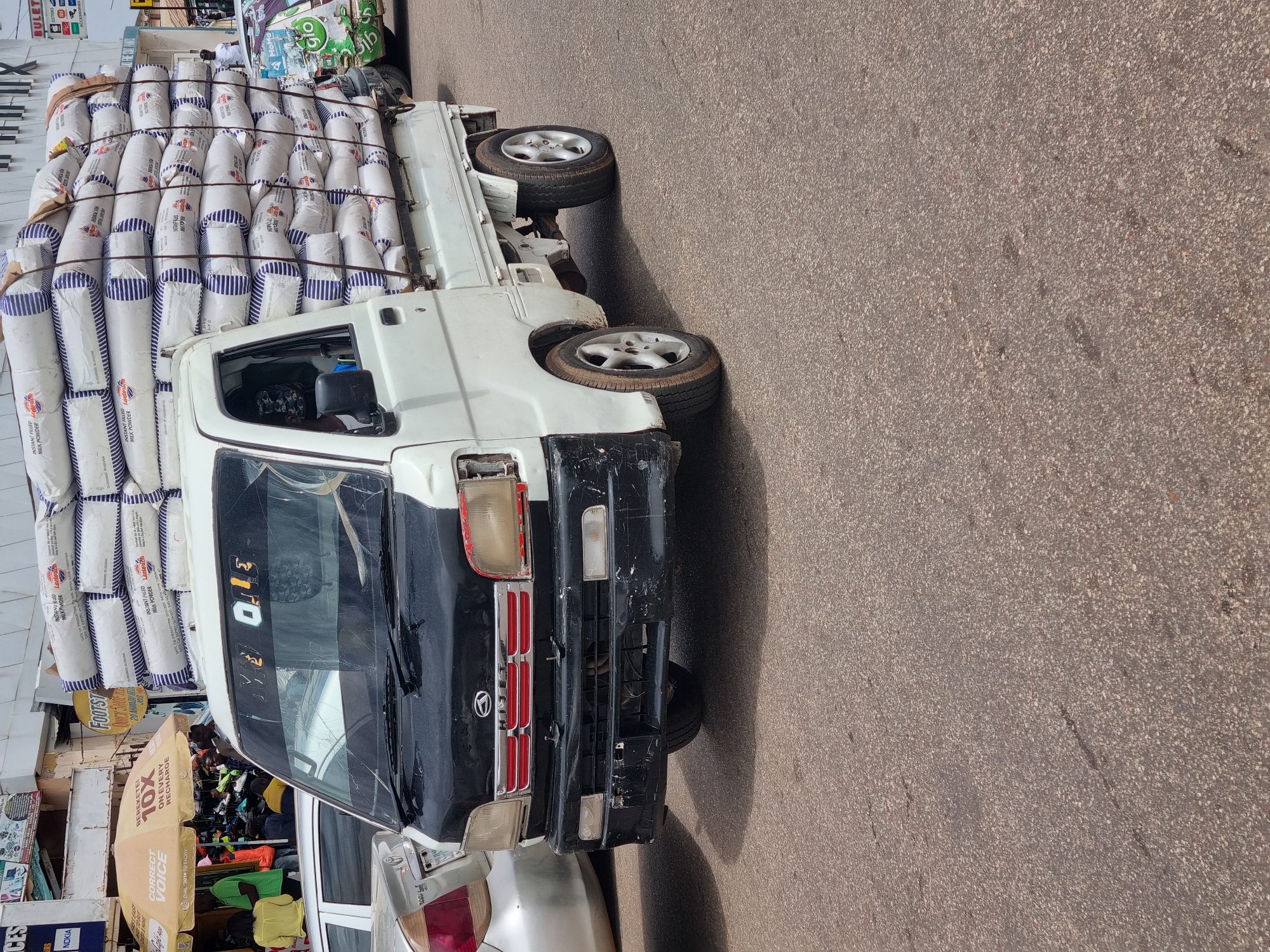 A photograph shows a small truck heavily loaded with bags of what appears to be powdered milk. The truck is white with a black front end and appears to be a Daihatsu. The windshield is cracked. The truck is parked on a paved road.
The truck bed is stacked high with bags labeled "Lactoch Instant Filled Milk Powder". The bags have a white body with blue stripes.
In the background on the left, there is a busy commercial area with people and shops. Signs for Nokia, Glo, and Footsteps can be seen. A sign advertising a promotion of "10X on every recharge" is also visible.
Based on the signage for "20 Ahmad Bello Way" and "Jos", the location is in Jos, Nigeria.