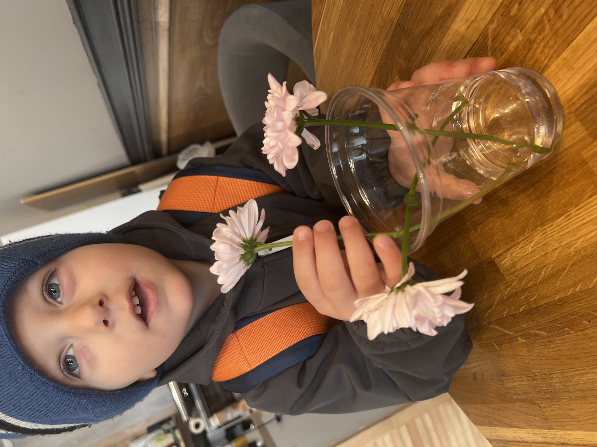 A young child with blue eyes and a blue hat is holding a clear plastic cup containing three light pink daisies. The cup is partially filled with water. The child is wearing a grey jacket with orange stripes on the sleeves. The background includes a wooden surface, a grey chair, and a glimpse of a window.
The location of the scene is indeterminate, as the city of Mytishchi, Russia cannot be confirmed without explicit supporting details in the image.