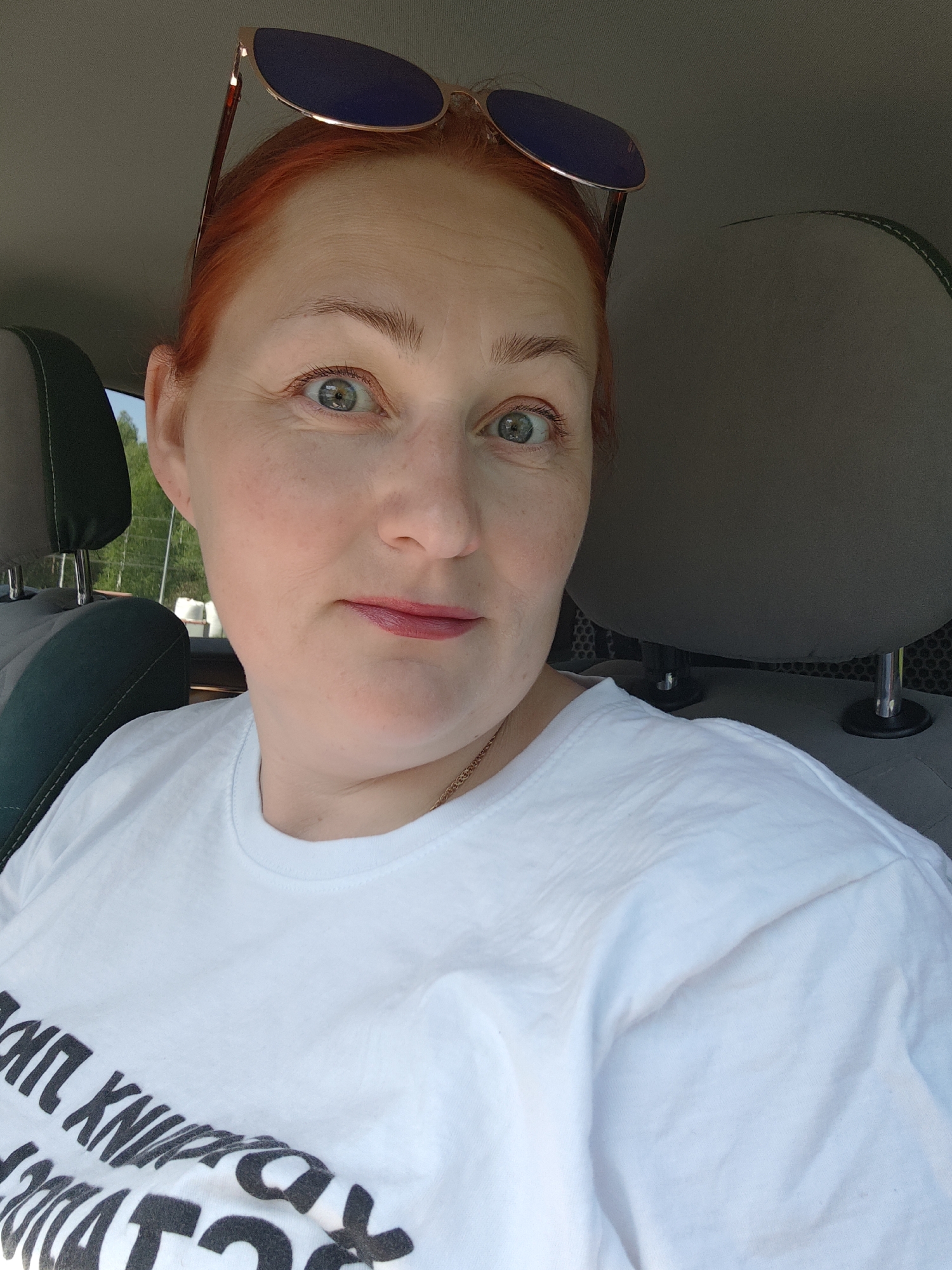 A red-haired woman with blue eyes is seated in a car, wearing a white T-shirt and sunglasses perched on her head. The car's interior includes gray seats with green accents and the vehicle appears to be parked in Chaikovsky, Russia, though this is not verifiable with this image.