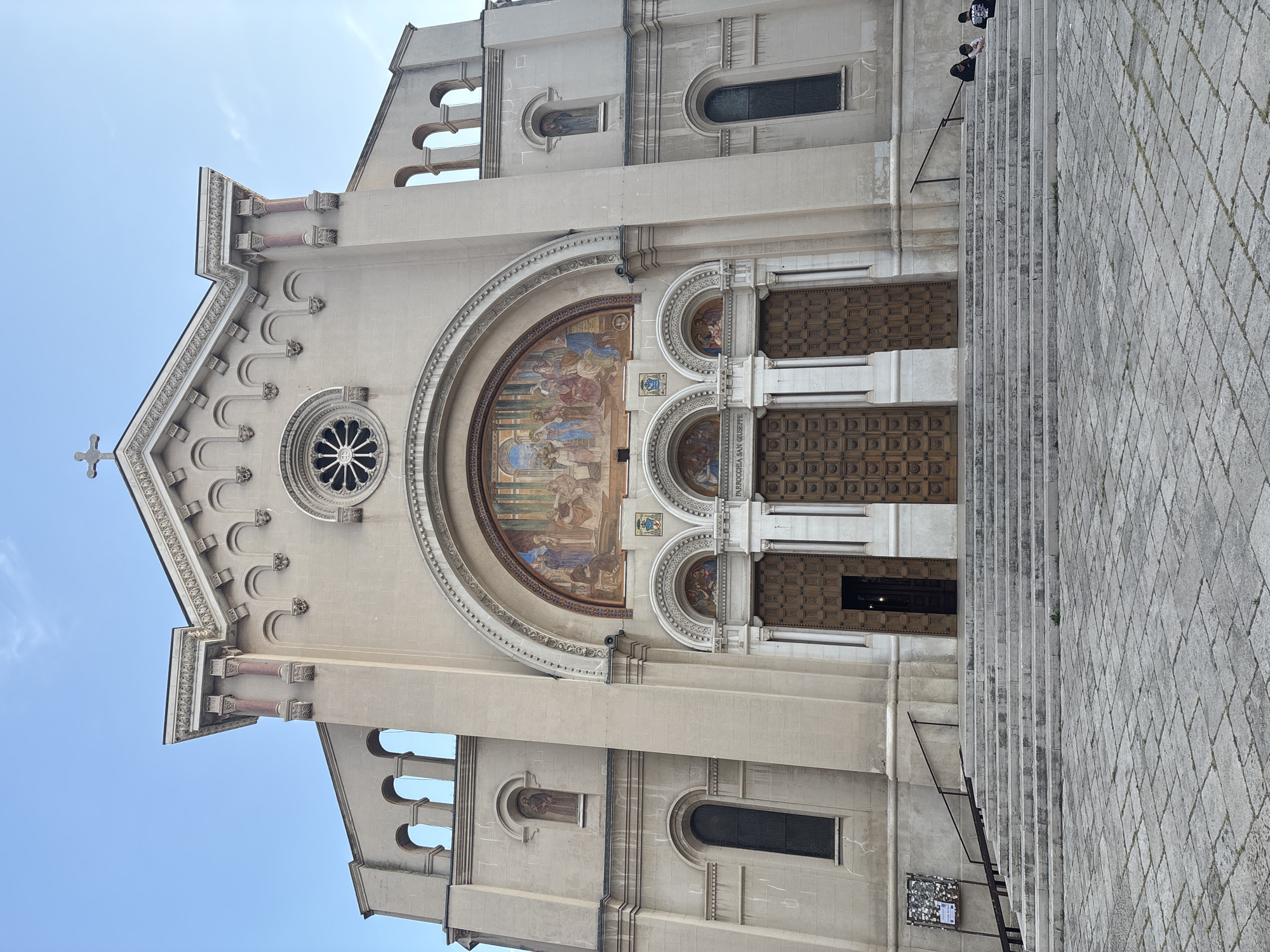 This image captures the grand facade of a light-colored church, appearing rotated 90 degrees counter-clockwise from its upright orientation. The central feature is a large arched entrance containing a colorful mosaic or fresco depicting a religious scene with multiple figures. Below this artwork are three tall, intricately carved wooden doors. Above the main arch, a circular rose window with a star-like design is prominent, and a simple cross crowns the peak of the gable roof.
The church's architecture is ornate, featuring decorative pilasters, cornices, and small arched windows and niches, some of which appear to hold smaller artistic details. The sky is clear blue with a few wispy clouds. To the right of the main entrance, a wide, stone staircase leads up to the building. One or two individuals, dressed in dark clothing, are visible on the upper steps of this staircase.
The scene depicts a daytime setting, likely morning or afternoon, with ample natural light illuminating the facade. A small sign is visible on an arched section above one of the side doors, displaying the text "PARROCCHIA SAN GIUSEPPE". This indicates the building is the Parish of Saint Joseph.