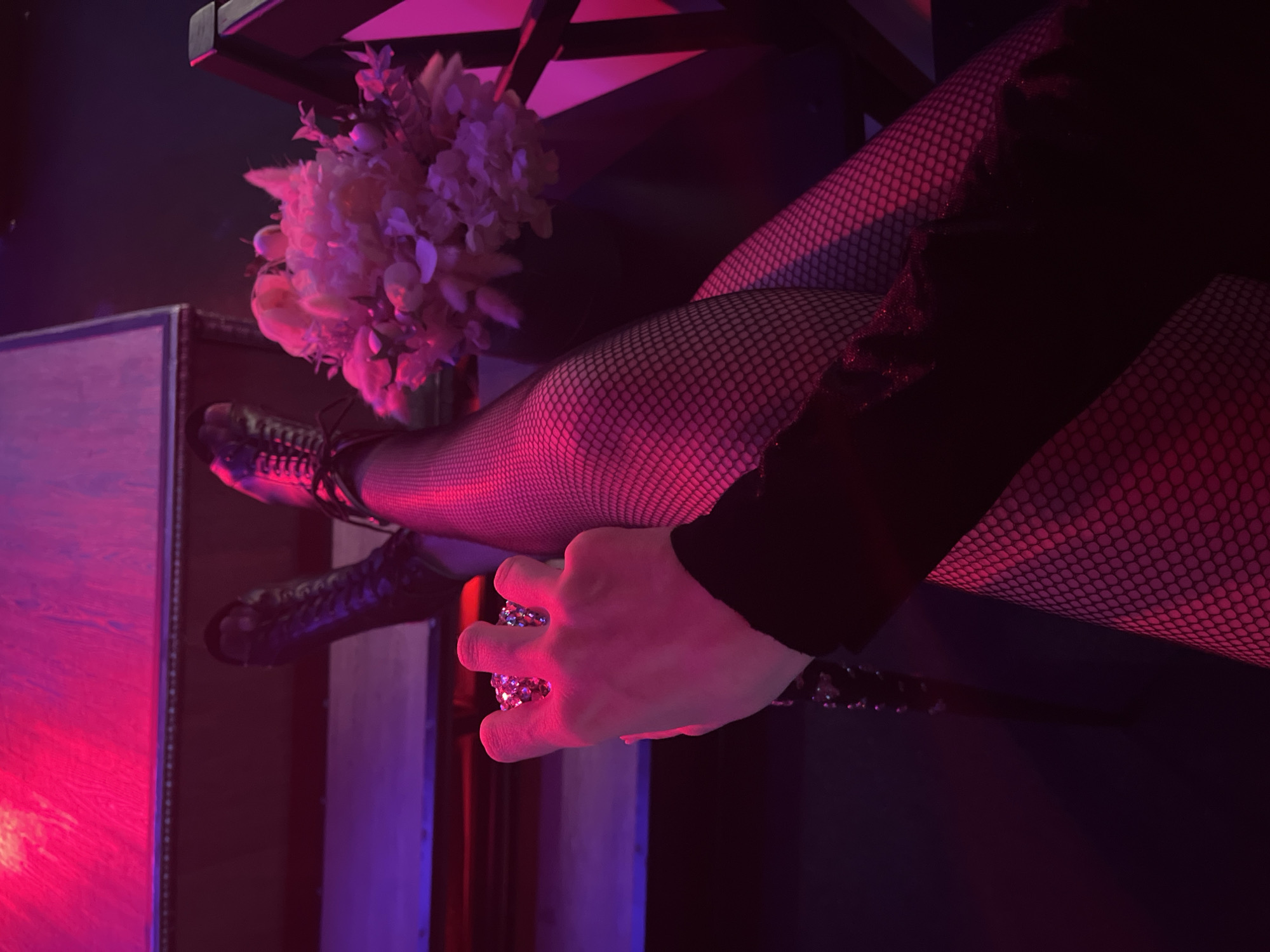 The image shows a person's legs and hand, illuminated by pink and red lights. The person is wearing black fishnet stockings and black lace-up boots. Their hand is holding a circular, glittery object. To the right, there is a bouquet of light pink flowers. The background suggests an indoor setting with steps or a tiered structure. The image does not contain any information that would indicate the location in the city of Kazan, Russia.