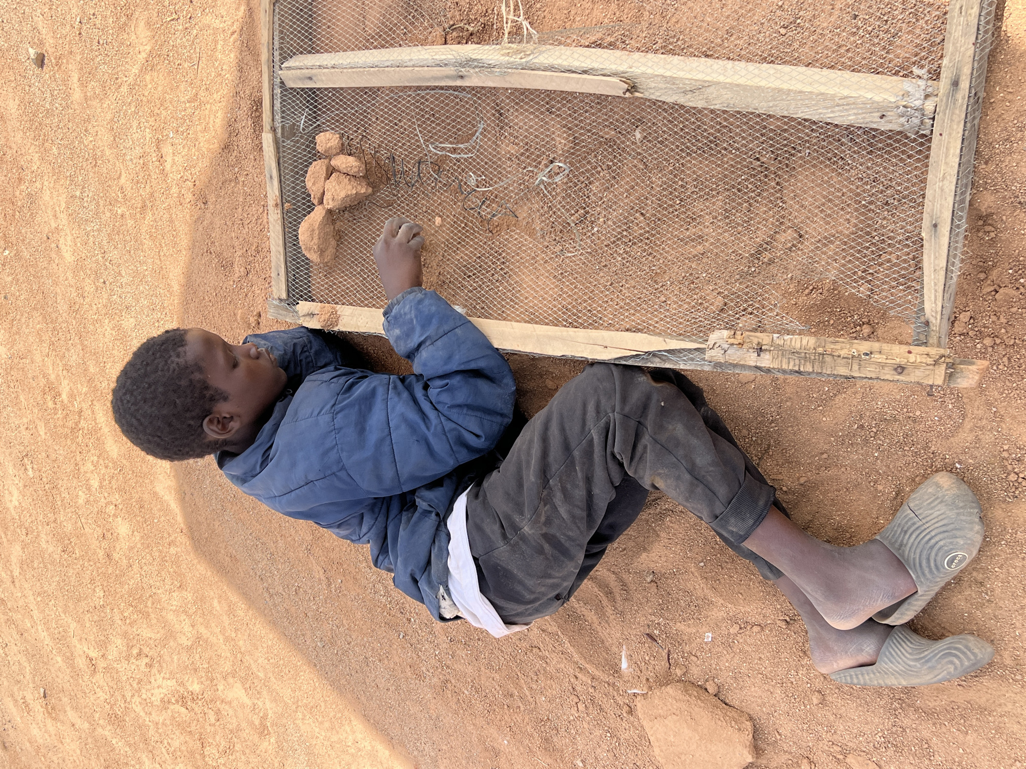 In Barakin Lerawi, Nigeria, a young male lies on the ground near a sieve constructed of wire mesh and a wooden frame. He is wearing a blue jacket, dark pants, a white undershirt, and gray Crocs-like footwear. The ground appears to be reddish-brown soil or sand. The boy is using his hand to manipulate objects, possibly stones or clumps of dirt, on the sieve. Pieces of wire are visible under the wire mesh.
