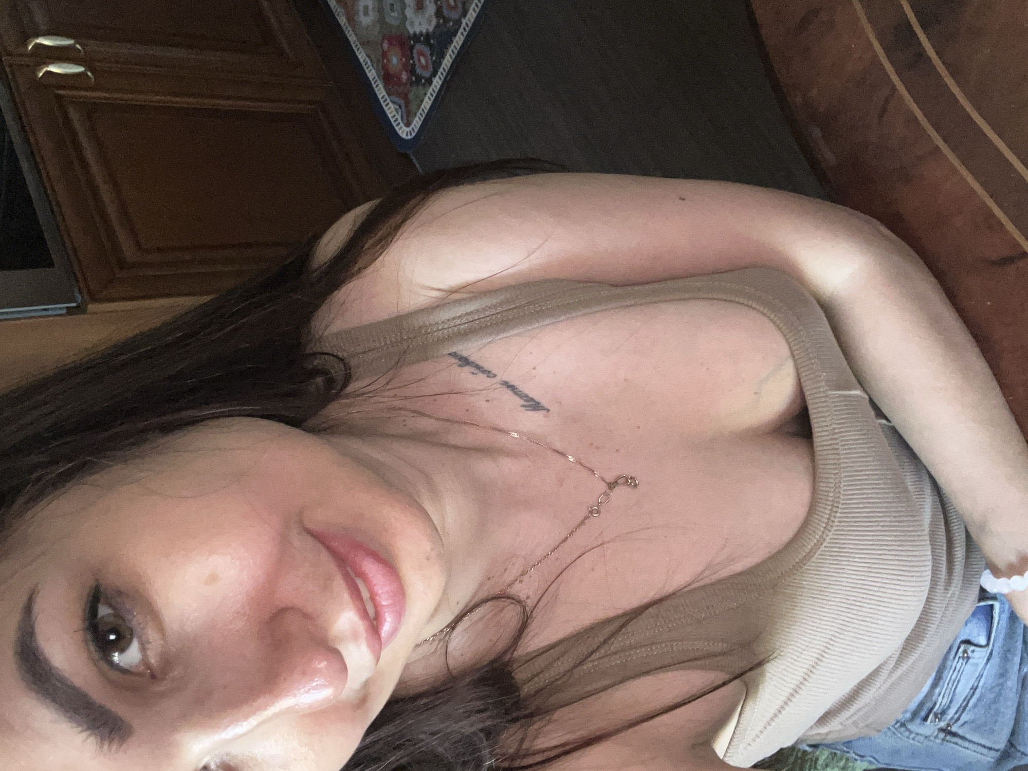 The image shows a close-up, selfie-style shot of a woman. She has long, dark hair, brown eyes, and fair skin. She is wearing a light brown, ribbed tank top and blue jeans. A delicate gold necklace with a small pendant is visible around her neck, and a subtle tattoo is on her collarbone. She is also wearing a white beaded bracelet on her right wrist. The woman is smiling softly, and her expression appears relaxed and content. The lighting suggests it might be daytime, though the exact time is unclear. The background is out of focus, but hints of wooden cabinetry and a patterned mat can be seen, suggesting an indoor domestic setting, possibly a kitchen or dining area. No specific event or activity is taking place; the image appears to be a candid or posed portrait.